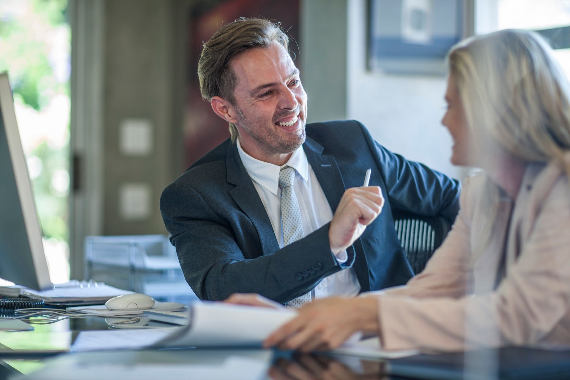 Man in a suit smiles and gestures, talking to a woman at a desk in an office setting.