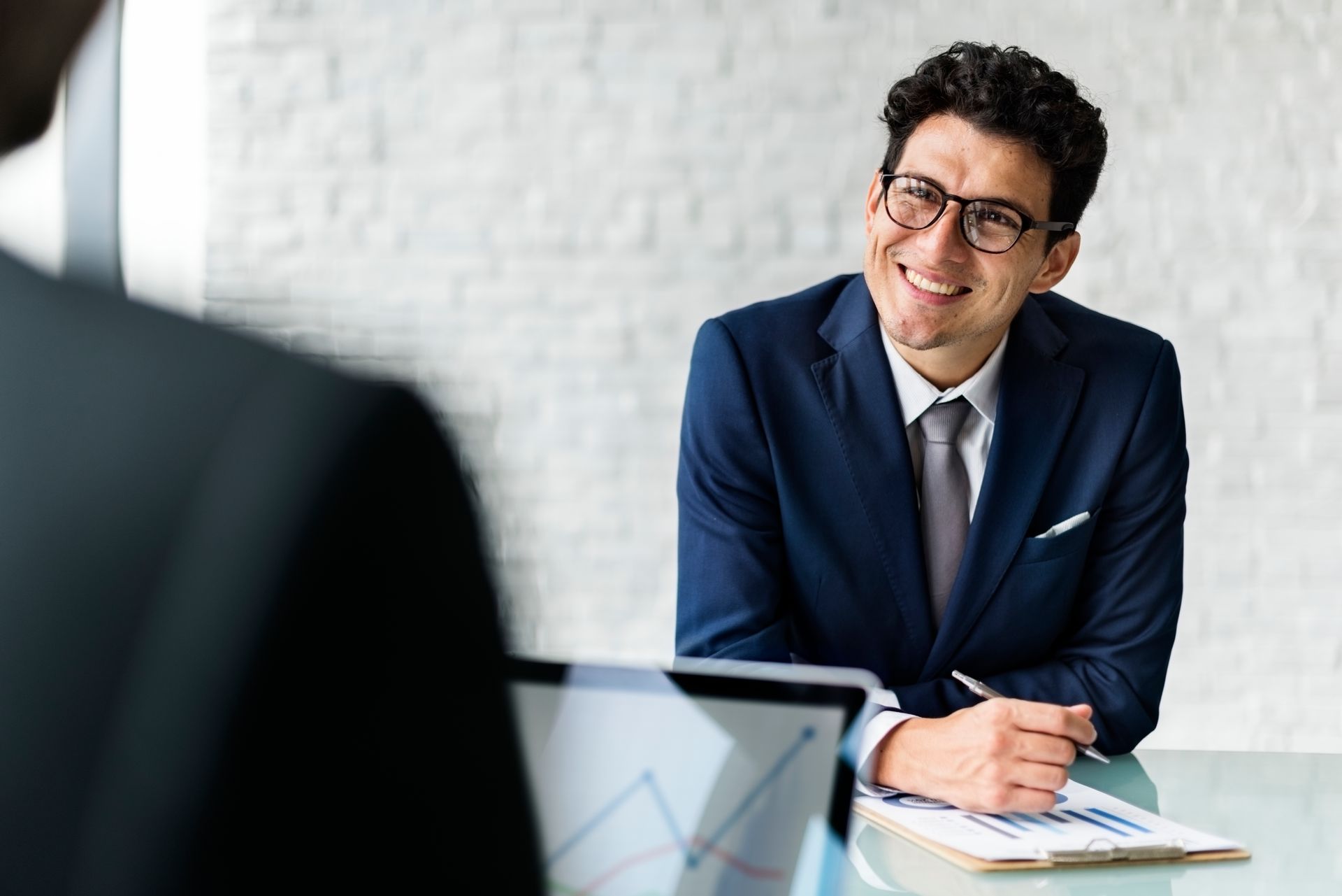 A smiling professional in a navy suit and glasses sits at a desk during a meeting, holding a pen over a document.