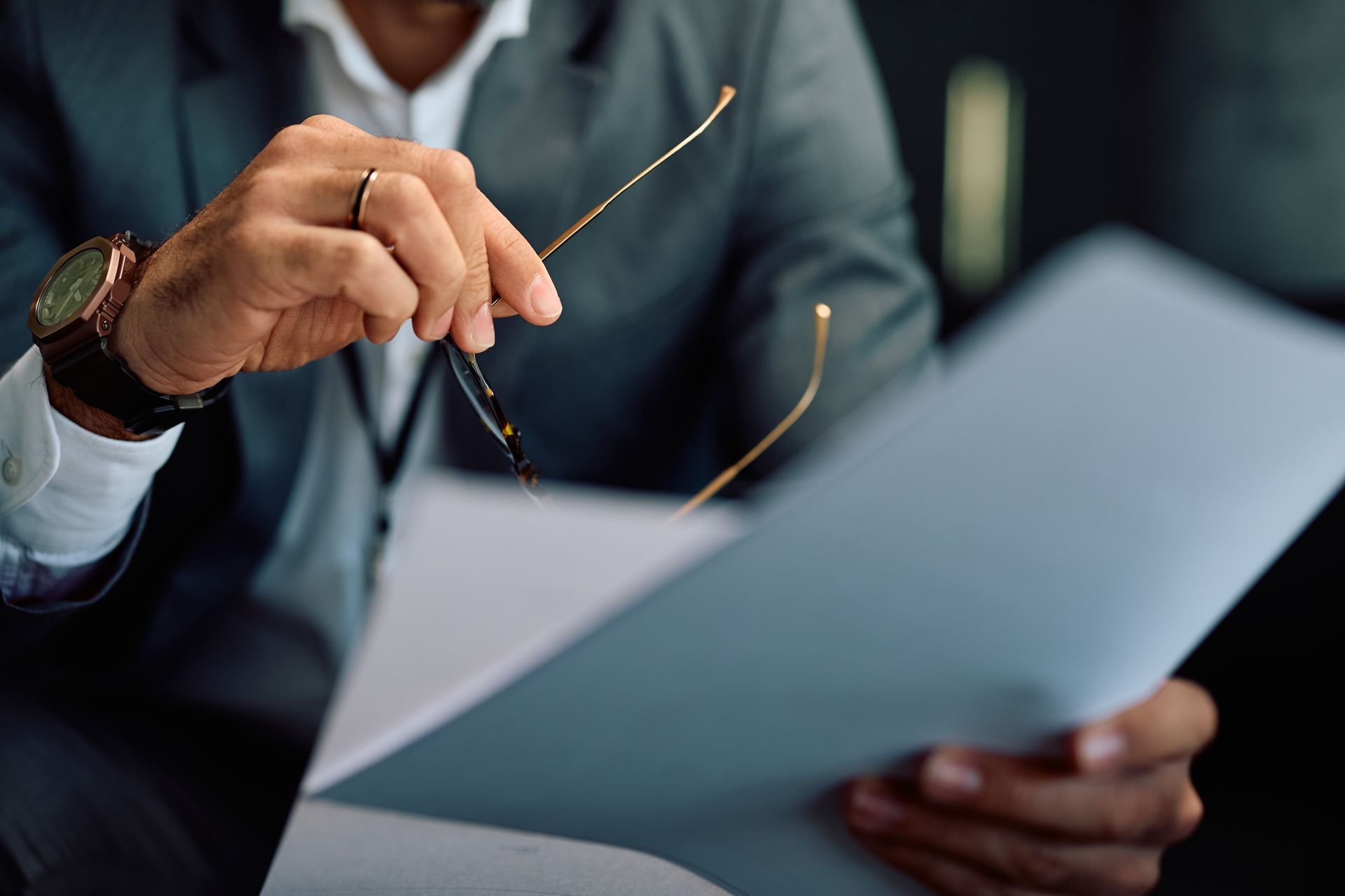 A person in a business suit holds a pair of glasses while reviewing a document.