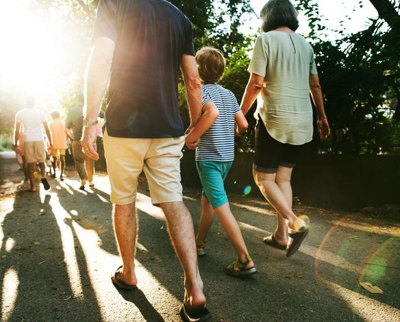 A family walks together on a sunlit outdoor path, viewed from behind.