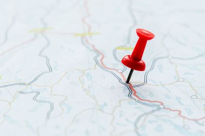 A bright red pushpin is stuck into a light-colored map showing winding roads and terrain.