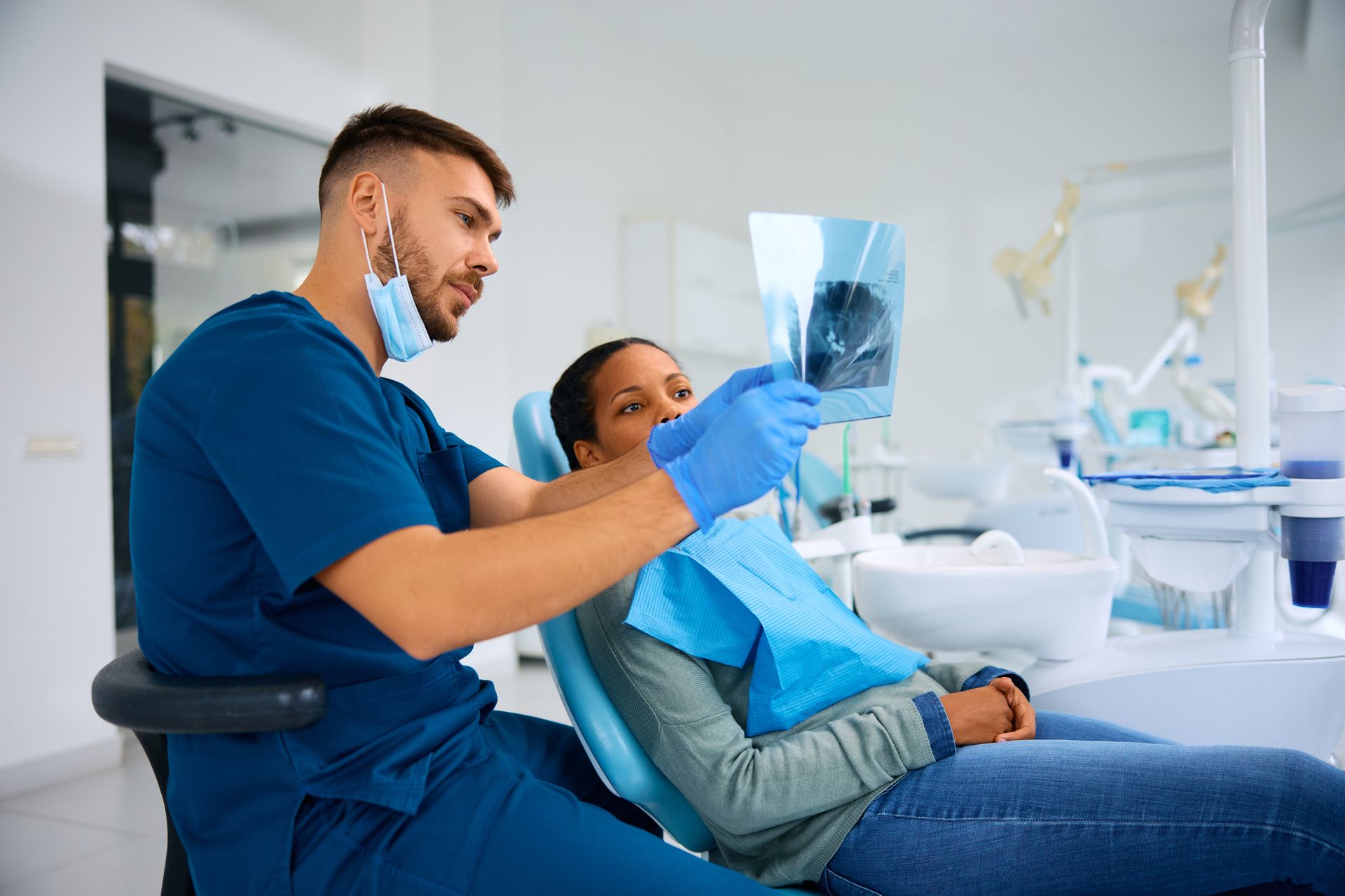 A dentist in blue scrubs and a face mask examines an X-ray film with a patient in a dental chair.