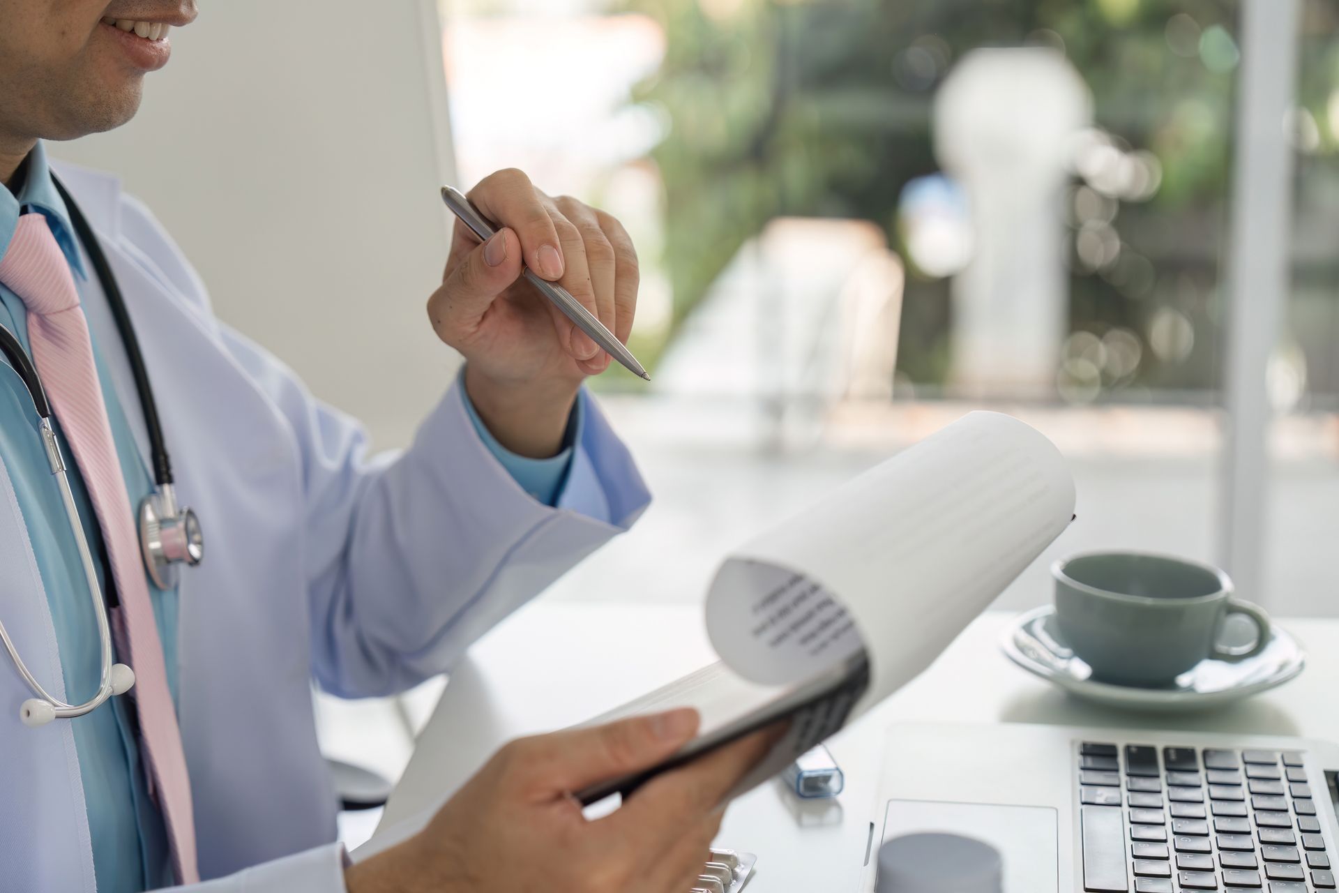 A medical professional in a white coat and pink tie holds a pen and a notepad at a desk with a coffee cup and laptop.