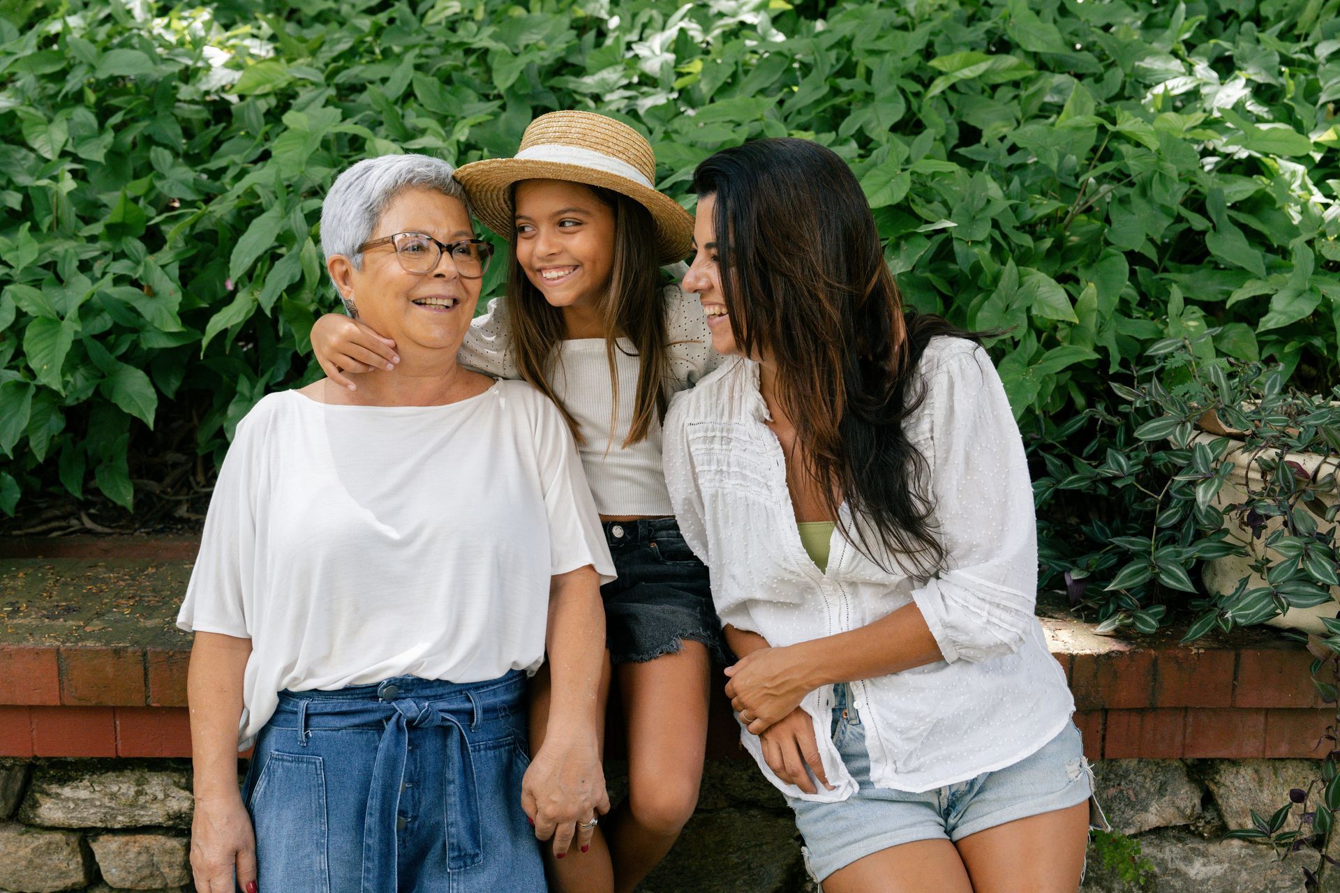 Three people smiling and posing together in front of a lush green hedge and a low brick wall.