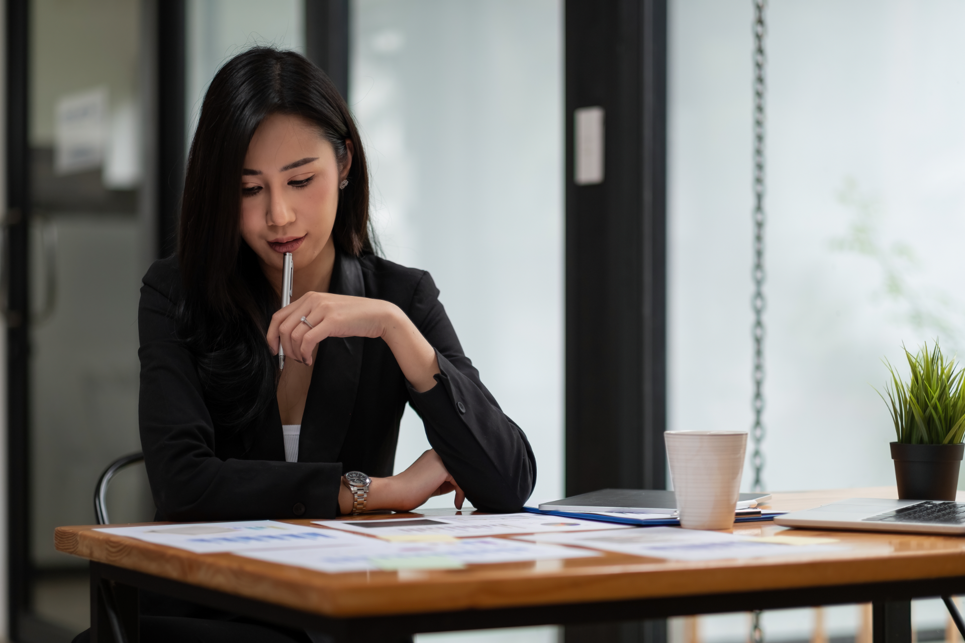 A person in a black blazer sits at a wooden office desk, thoughtfully holding a pen while reviewing documents.
