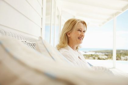 A person with blonde hair smiles while relaxing on a white lounge chair on a bright, outdoor porch overlooking the water.