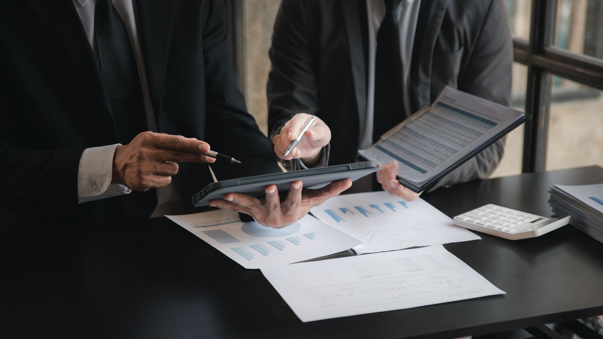 Two people in business suits analyze financial charts and a digital tablet at a desk.