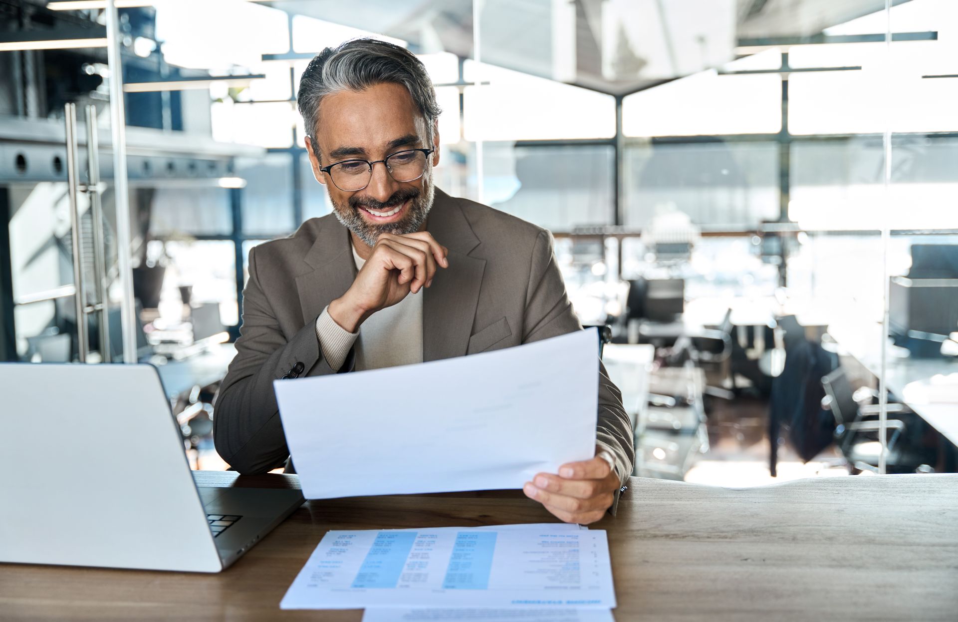A professional sits at a desk with a laptop and documents, smiling while reviewing paperwork in a modern office.