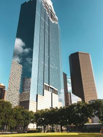 A tall, blue, reflective skyscraper towers over other buildings and trees in a sunny urban park setting.