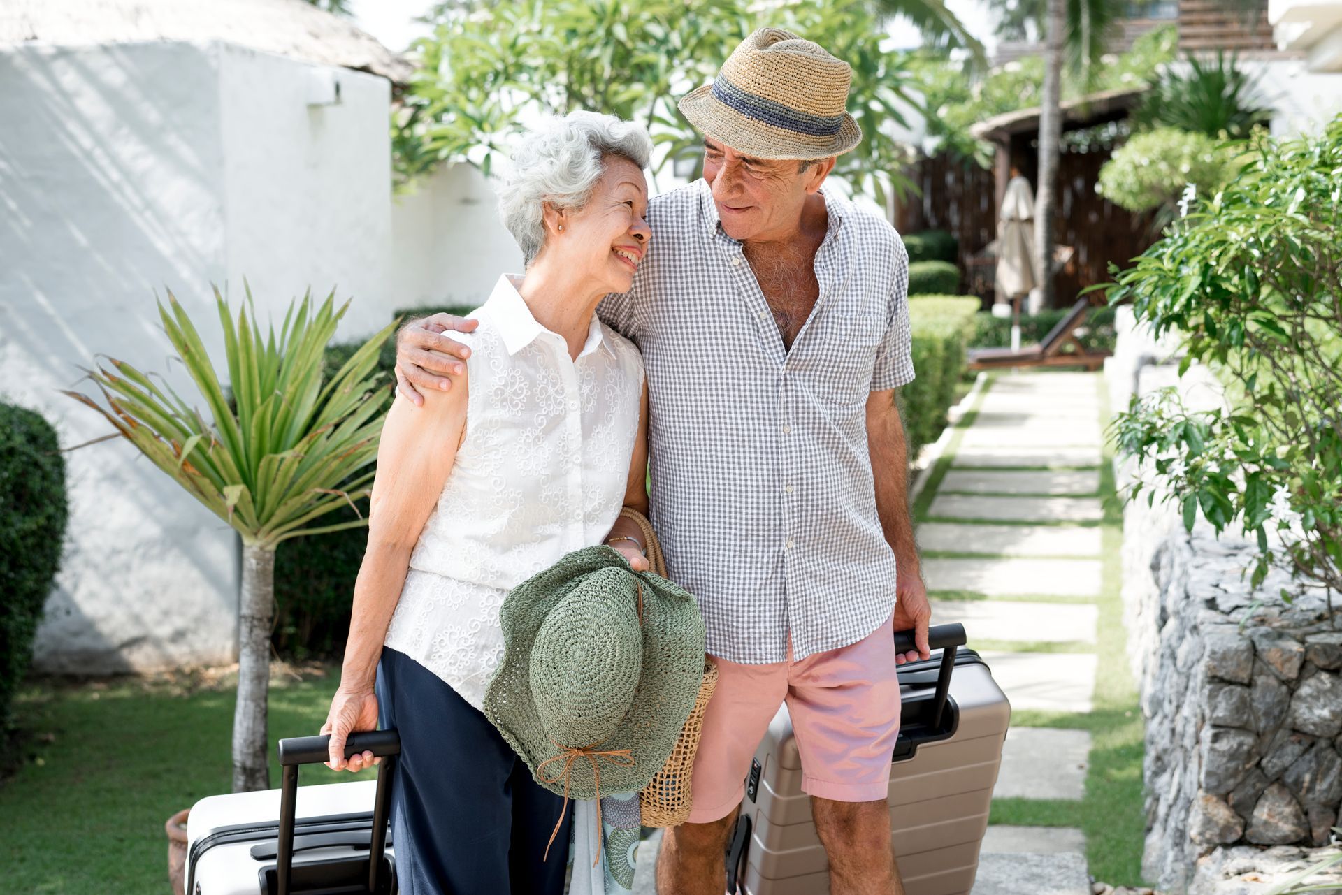 A couple in casual vacation attire walks together on a path while pulling their suitcases outdoors.