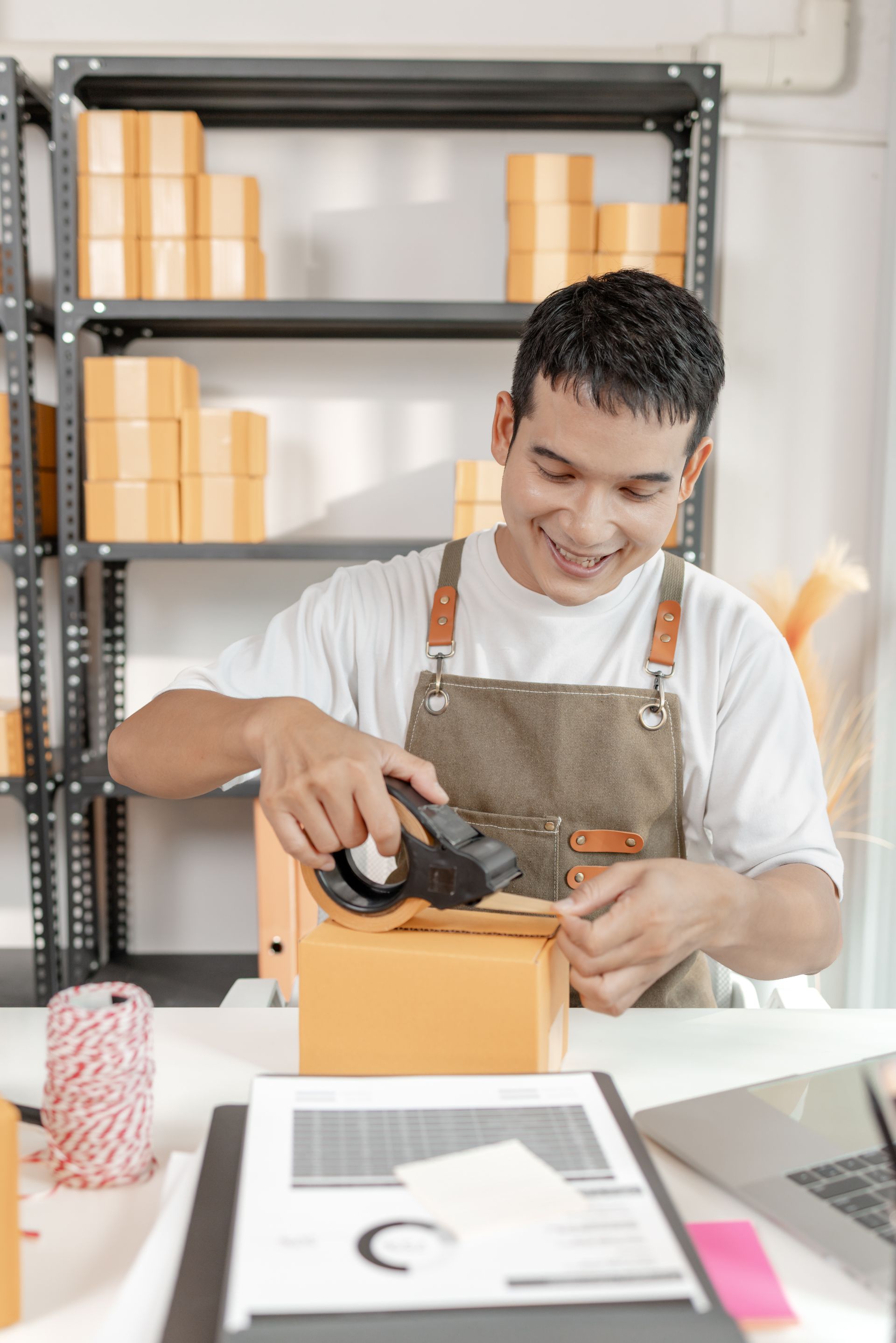 A person wearing an apron smiles while sealing a cardboard shipping box with a tape dispenser in a storage room.