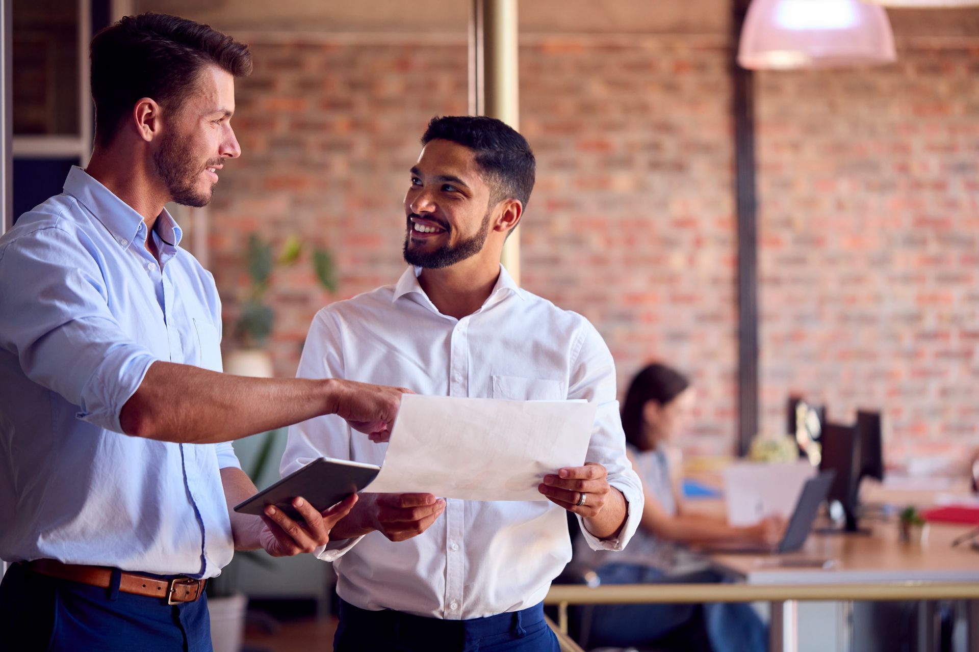 Two professionals in light-colored shirts reviewing documents and a tablet together in a modern, brick-walled office.