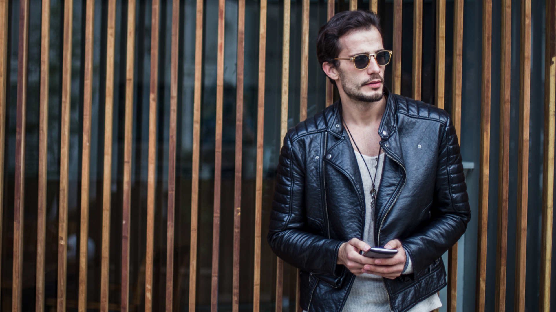 A man in a leather jacket and sunglasses is standing in front of a wooden fence holding a cell phone.