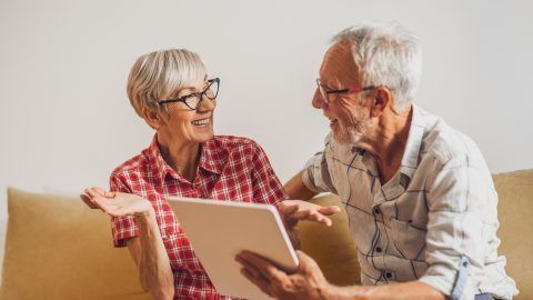 An elderly couple is sitting on a couch looking at a tablet.