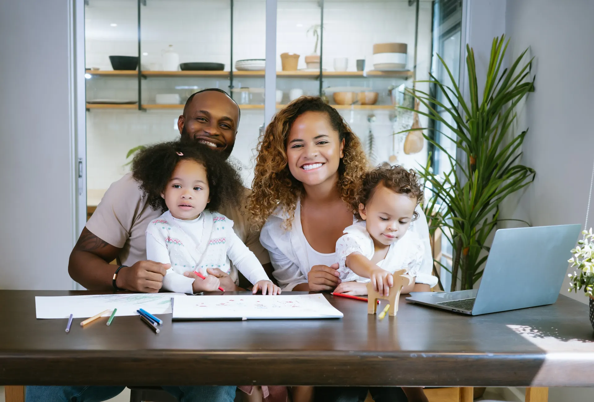 Family of four, including parents and two young children, smiles at a table with a laptop and coloring supplies.