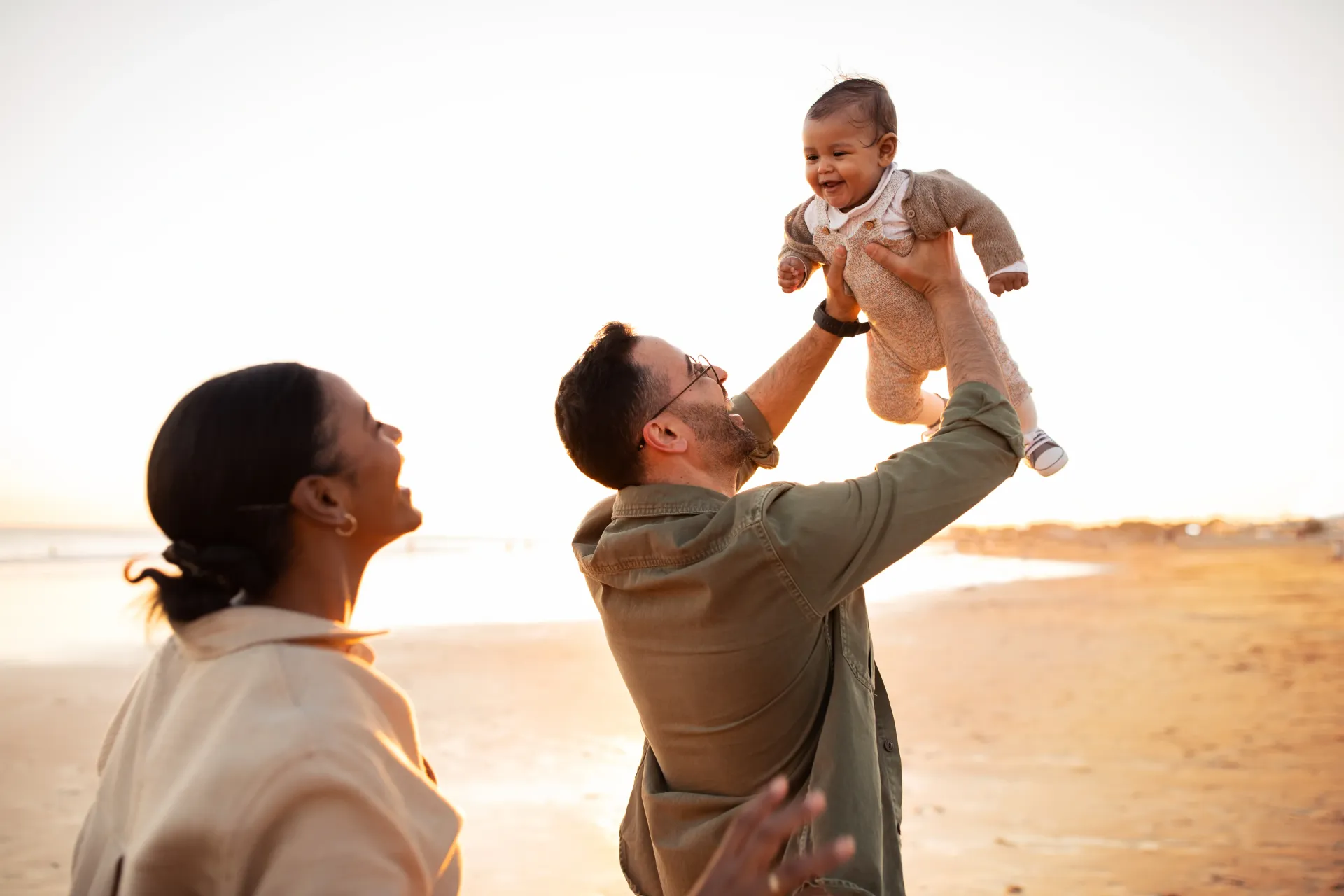 Parents on beach toss baby in the air; sunset illuminates family, evoking joy.