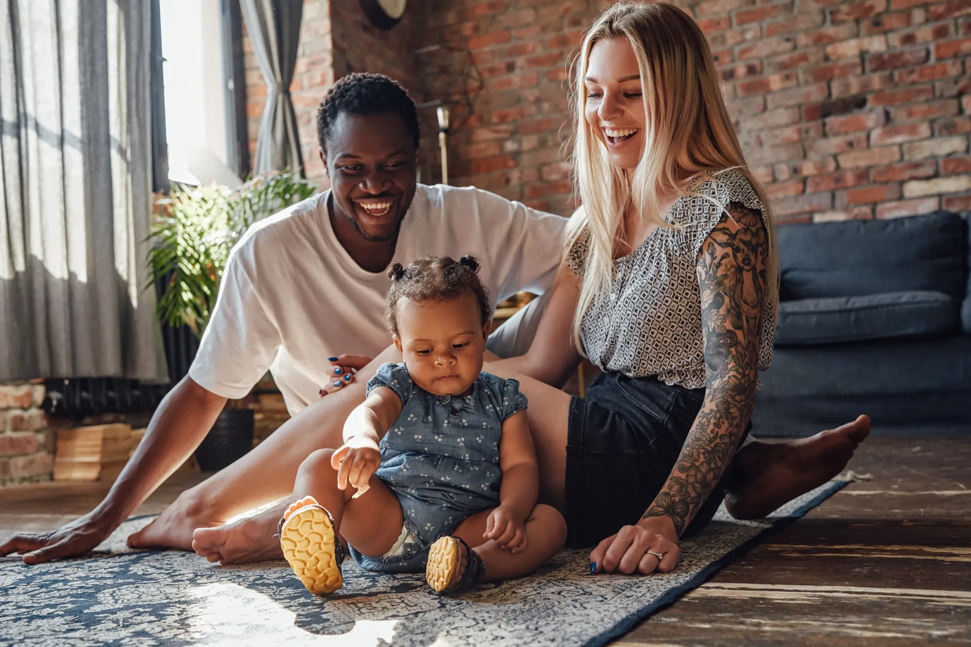 Family of three, Black father, white mother, and biracial baby girl, laughing together on a rug.