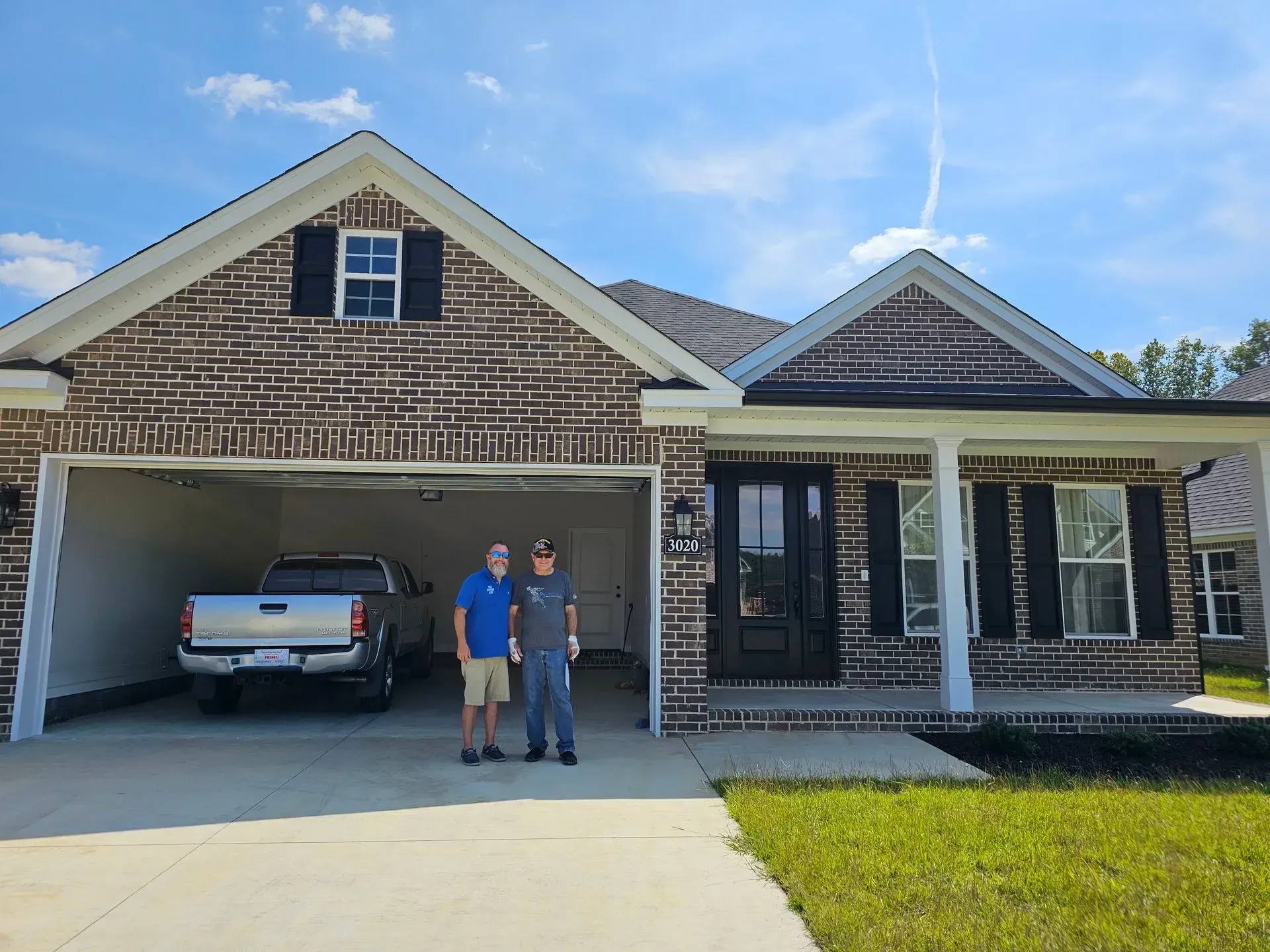 Two men are standing in front of a brick house with a truck parked in the driveway.