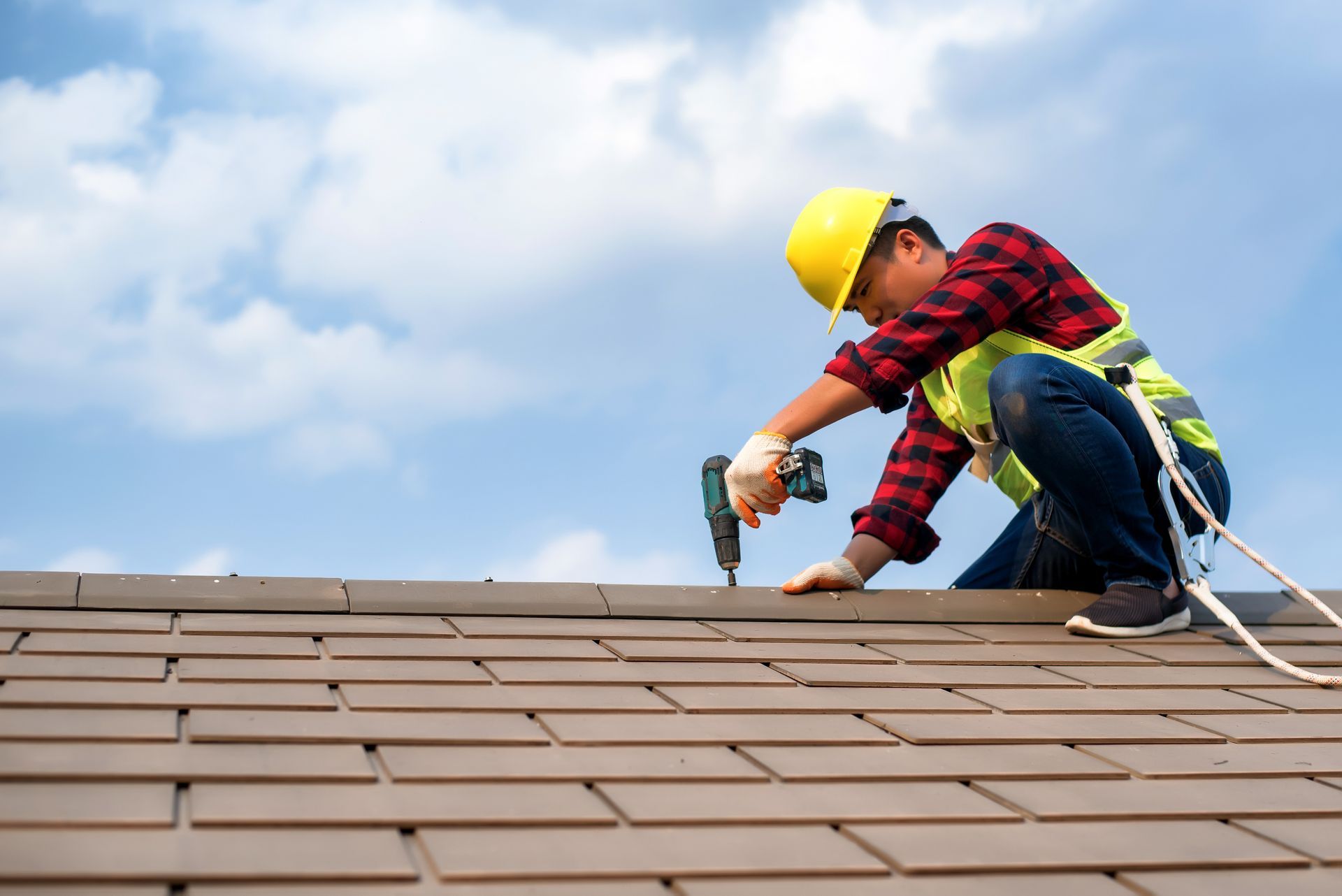 Worker in yellow hard hat repairing roof with a hammer under a blue sky