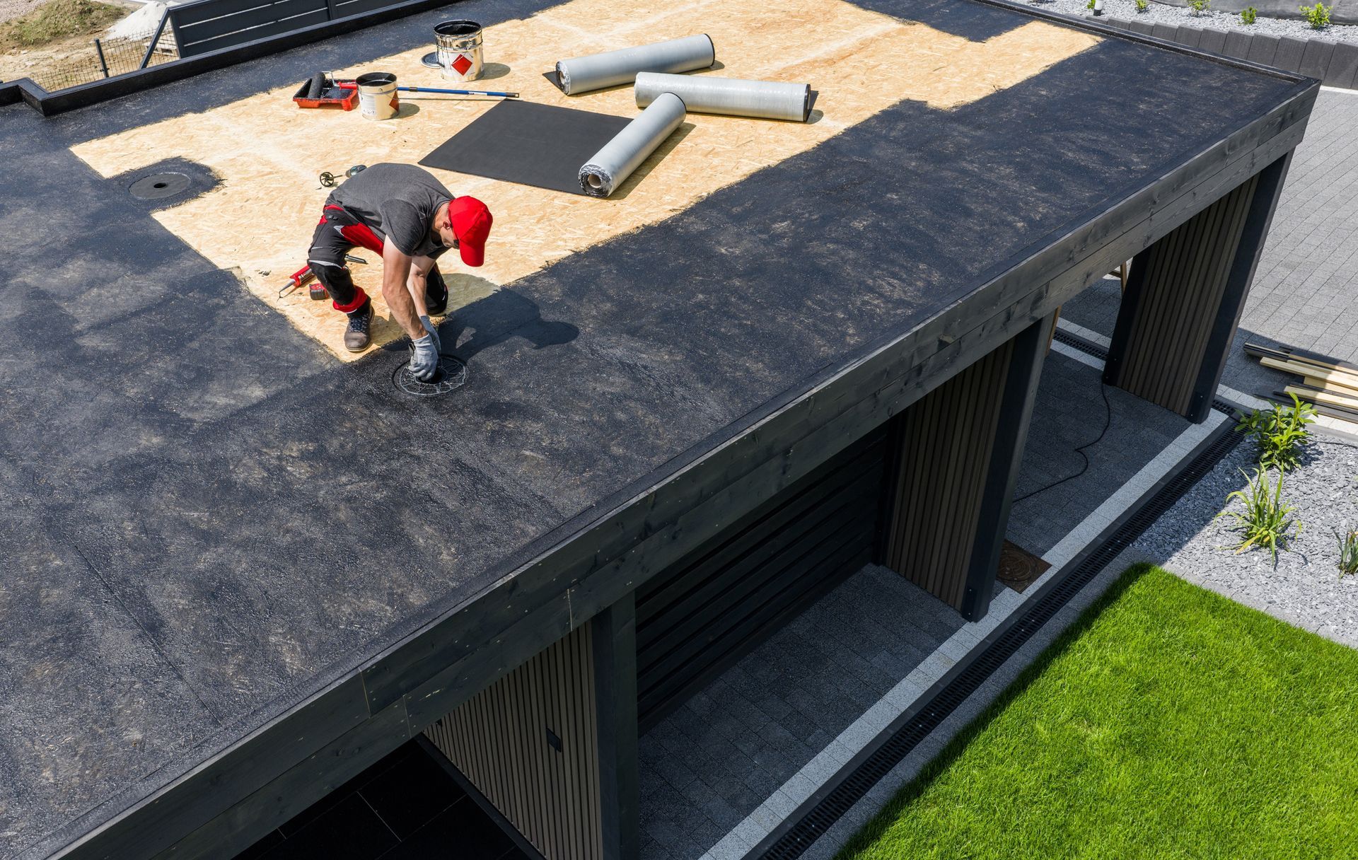 Worker on a rooftop installing materials on a flat roof next to pipes and a gravel area.