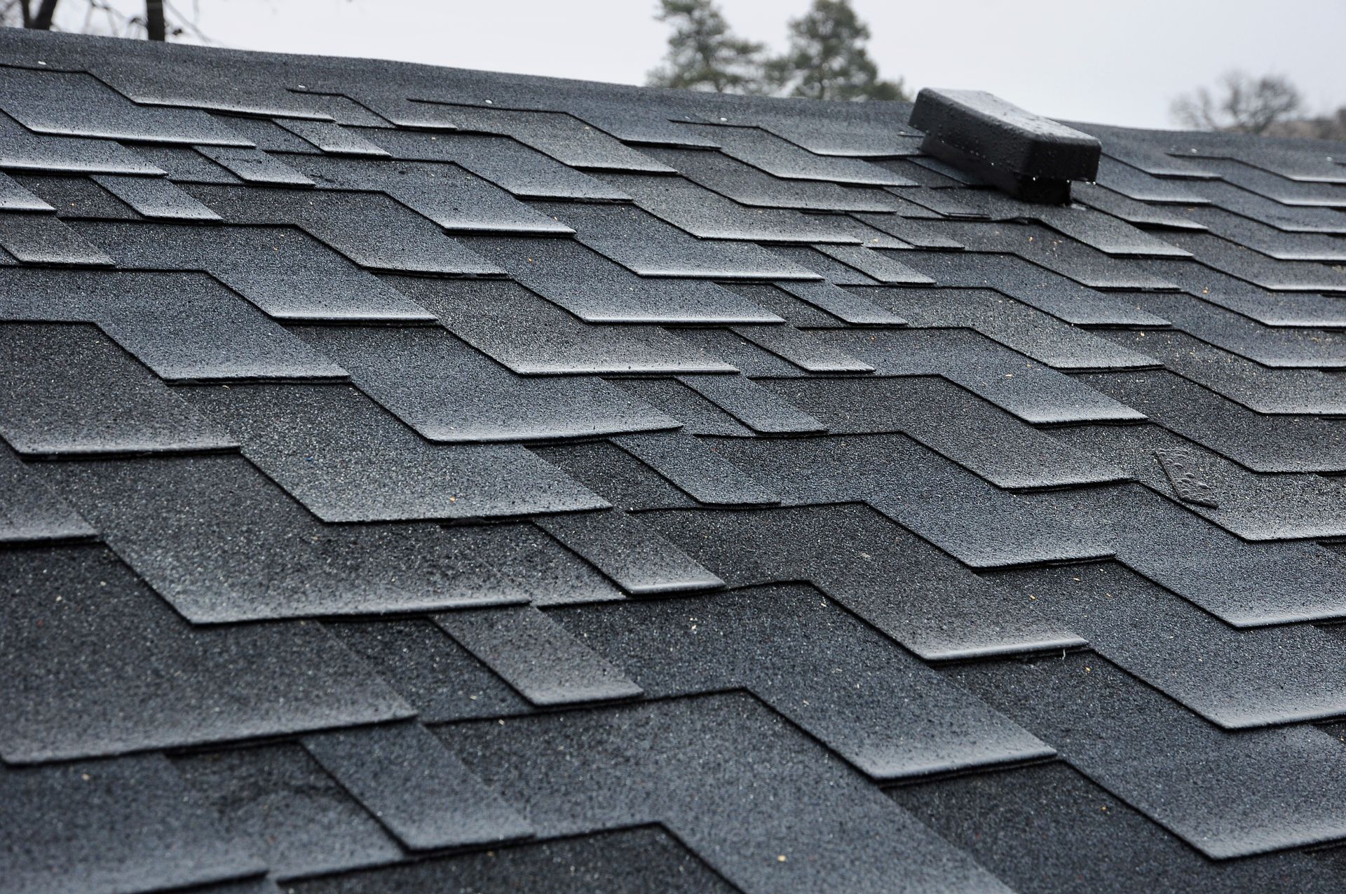 Dark gray asphalt shingles on a roof with a small vent against a cloudy sky
