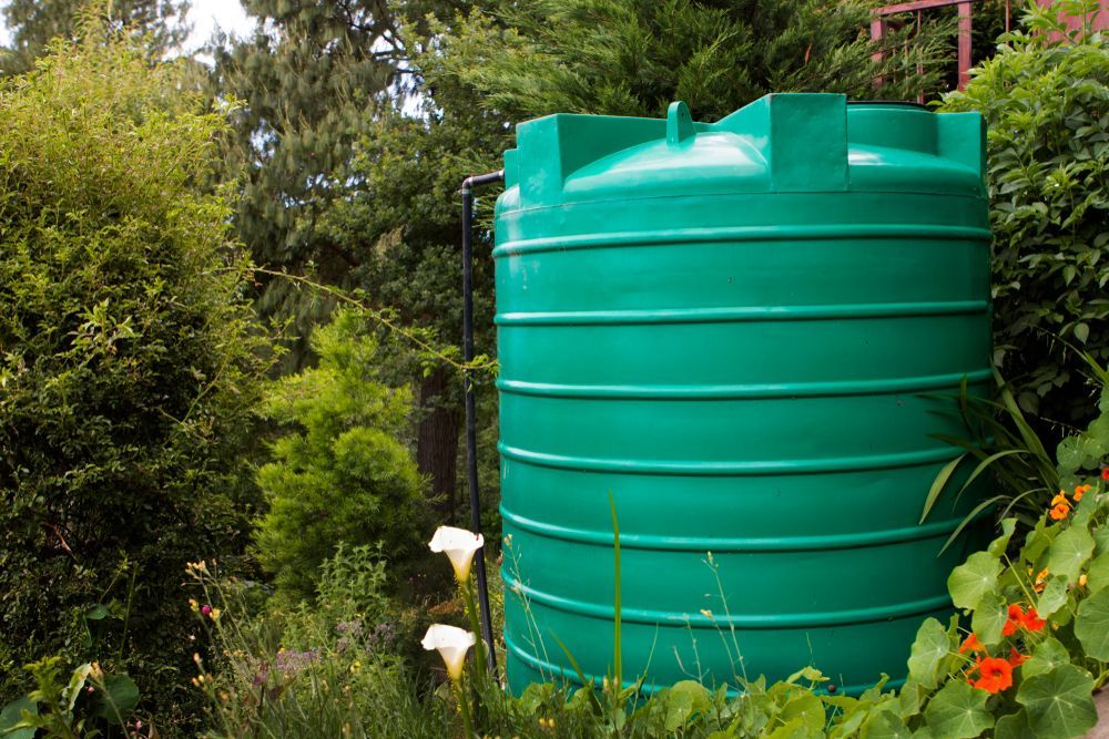 A Green Water Tank is Surrounded by Flowers and Trees in a Garden — Dave's Plumbing and Gasfitting in Maroochydore, QLD