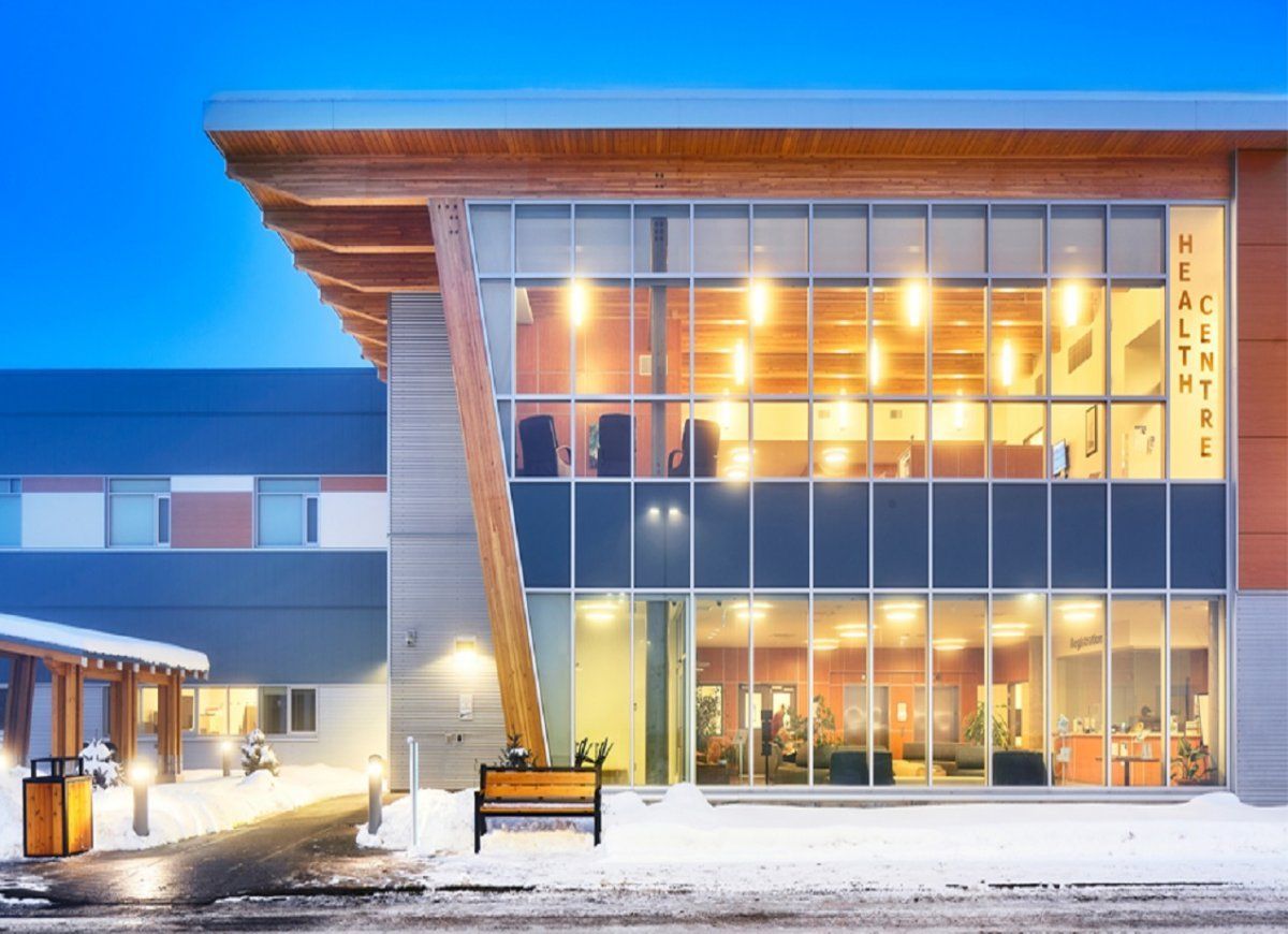 Modern health center exterior at dusk, snow covered ground, large windows, wood accents, illuminated interior.