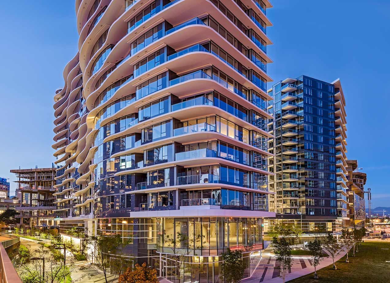 Modern high-rise residential buildings with curved balconies, glass facade, and illuminated base at dusk.