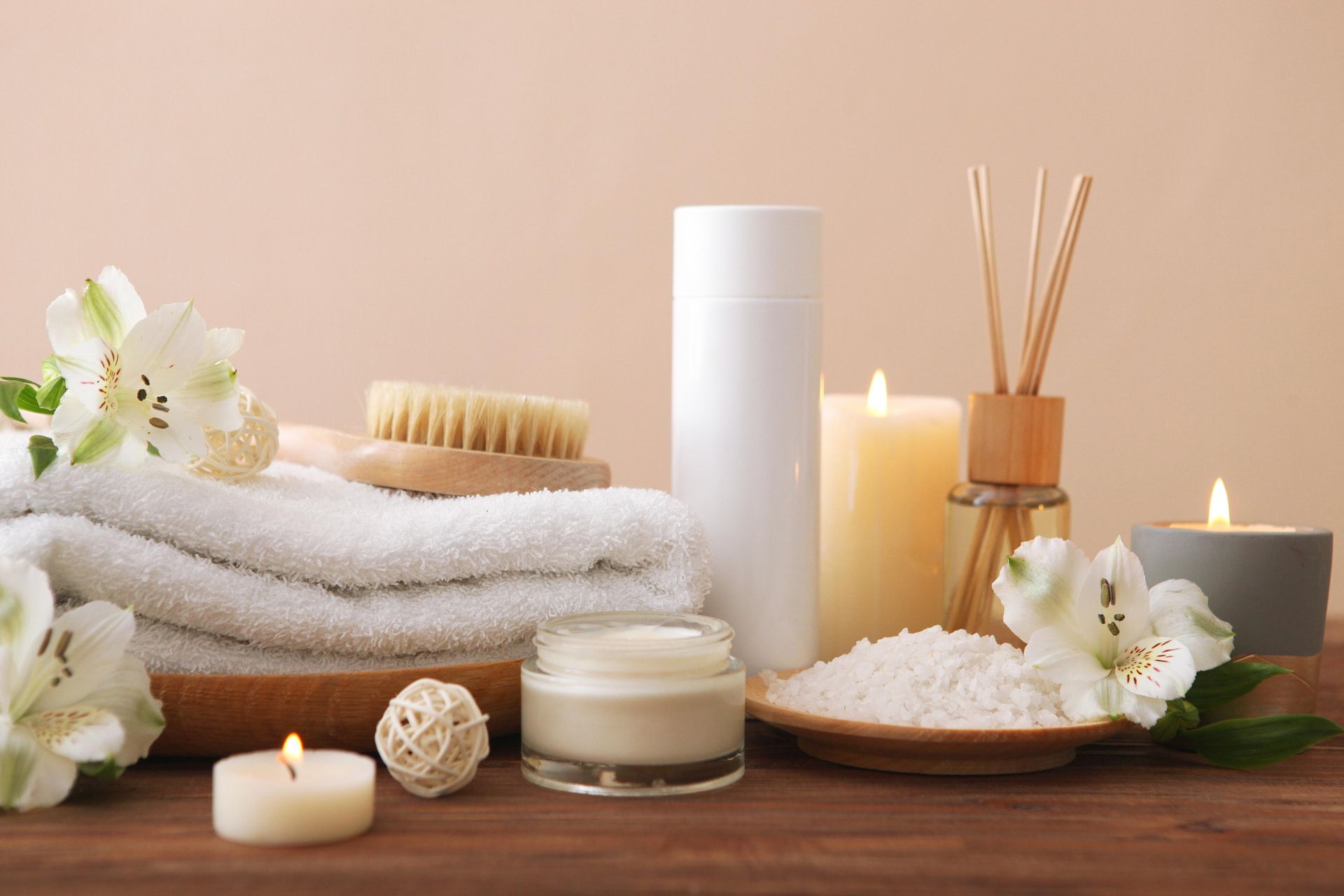 Spa scene: Stacked white towels, candles, flowers, beauty products on wood table, beige background.