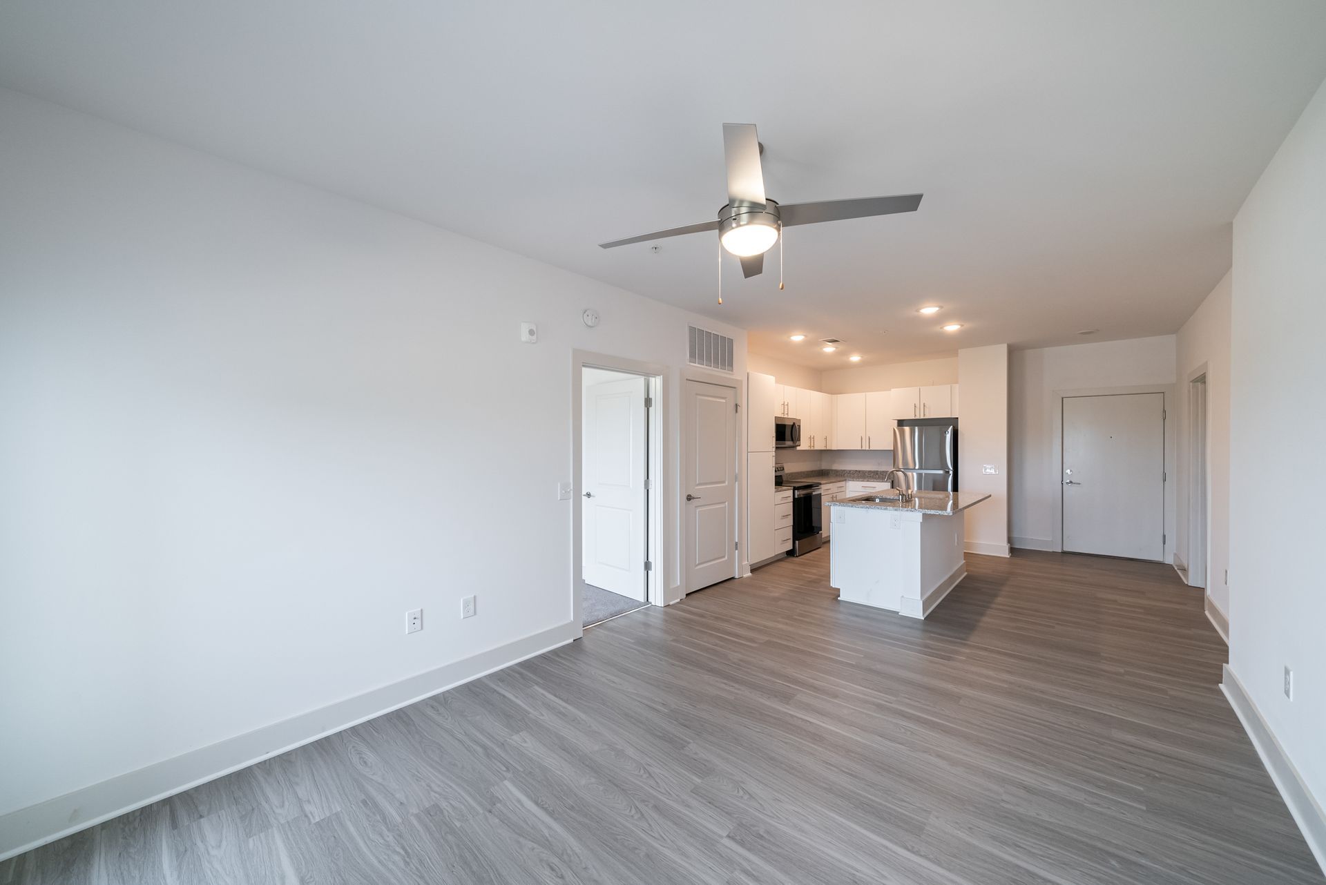 Empty apartment interior with light gray flooring, white walls, and open kitchen.