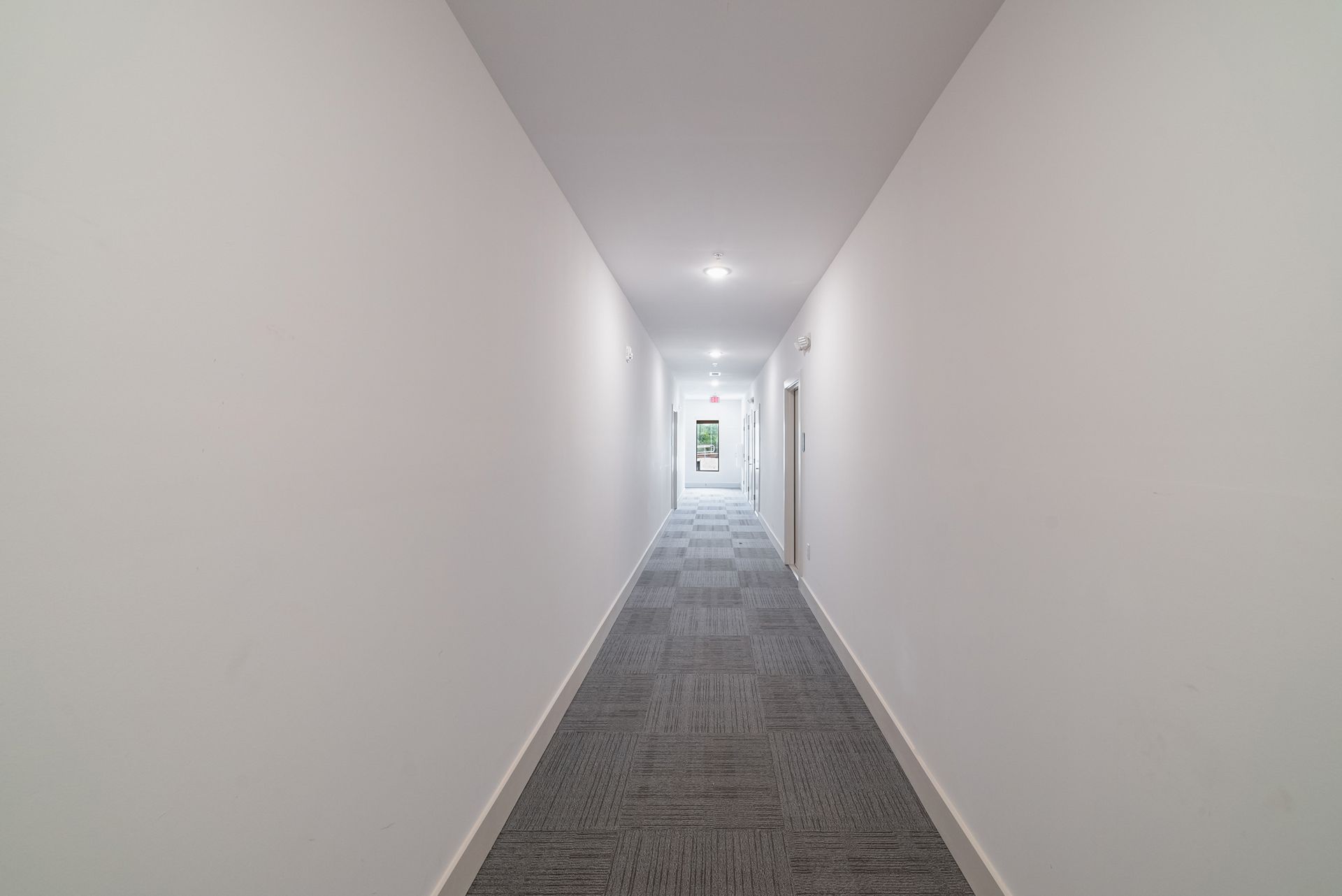 Long, narrow hallway with white walls, gray carpet, and recessed lighting.