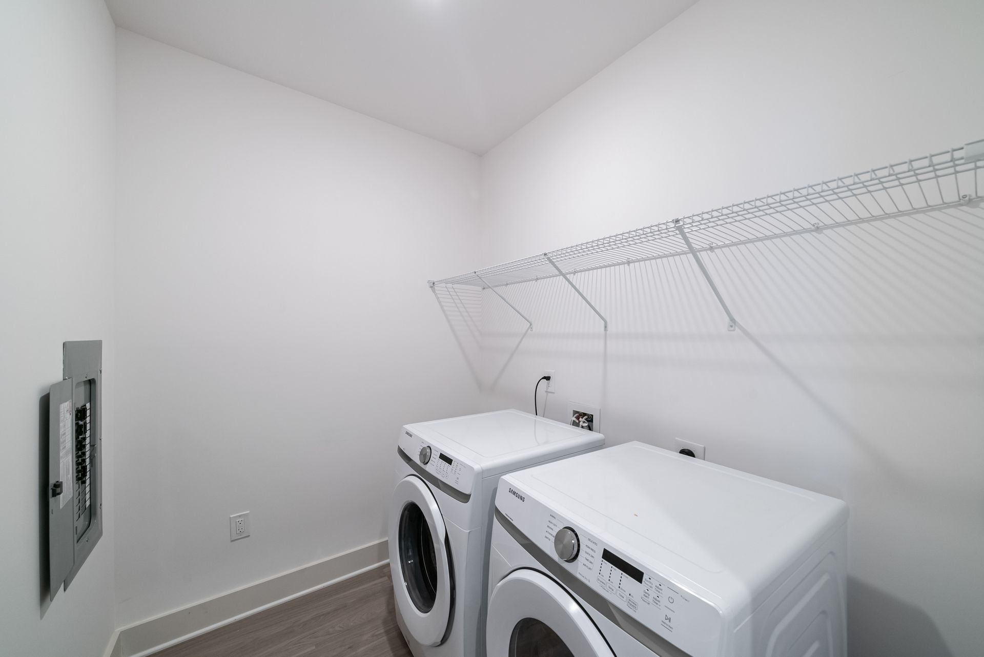 Laundry room with white washer and dryer under a wire shelf. A grey electrical panel is on the wall.