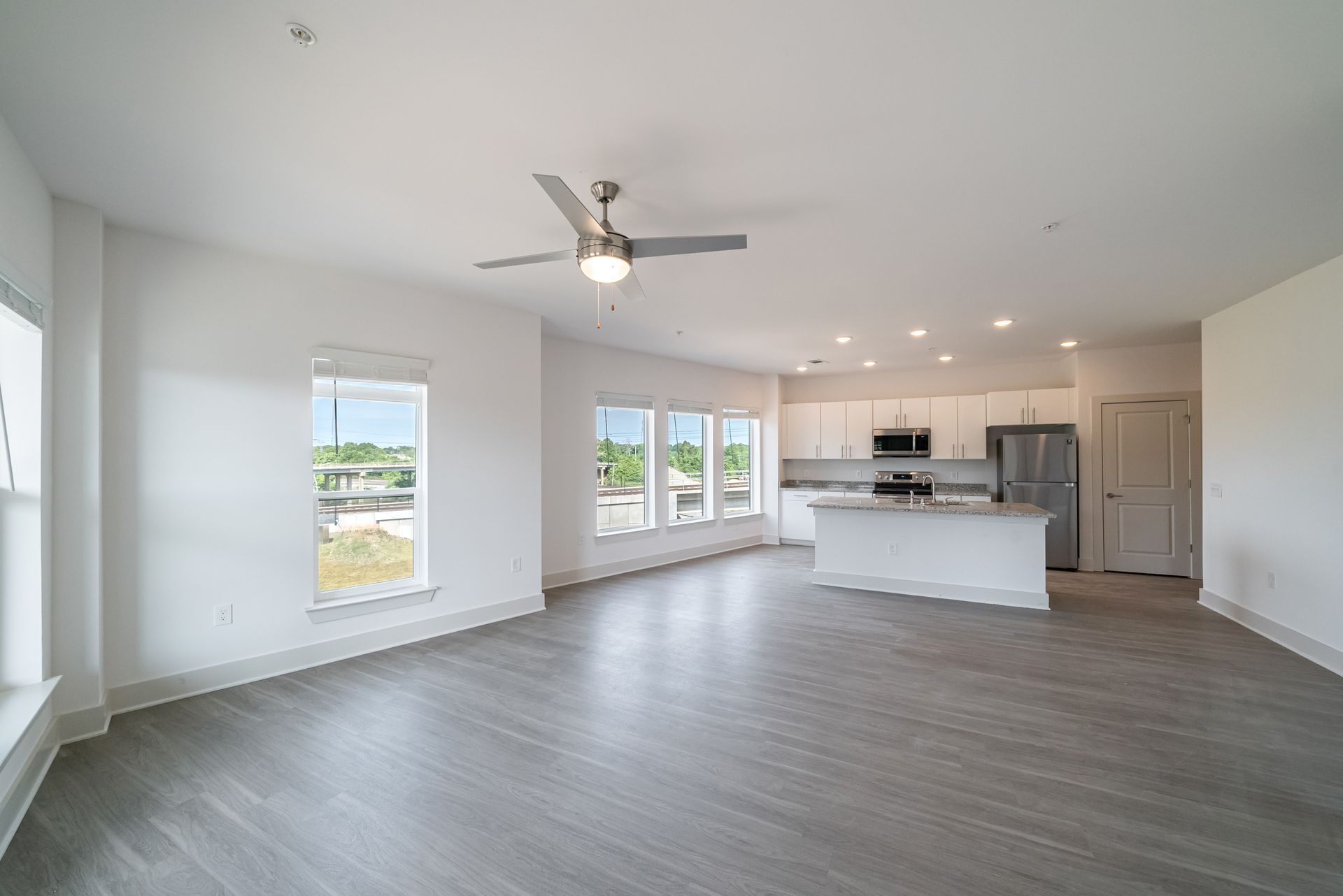 Empty apartment with open kitchen; white walls, gray floors, windows, and ceiling fan.