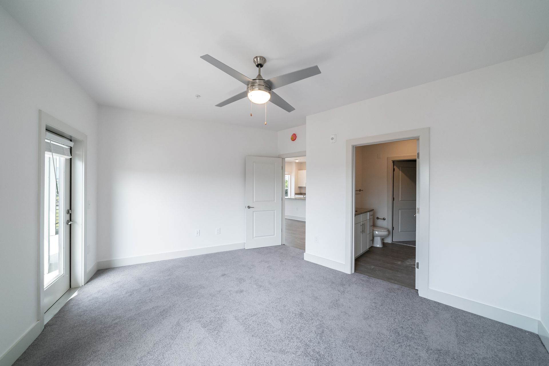 Bedroom with gray carpet, white walls, door to bathroom, ceiling fan, and open door to balcony.