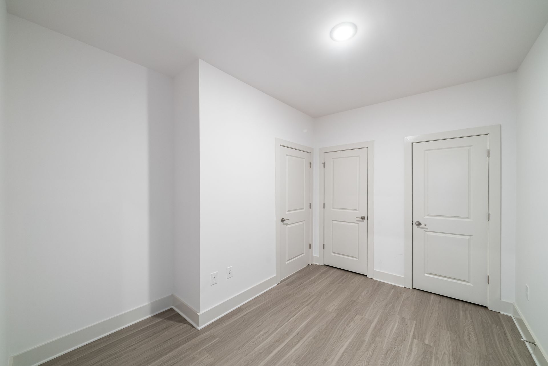 Empty, white room with three doors and light gray flooring; the ceiling has a single overhead light.