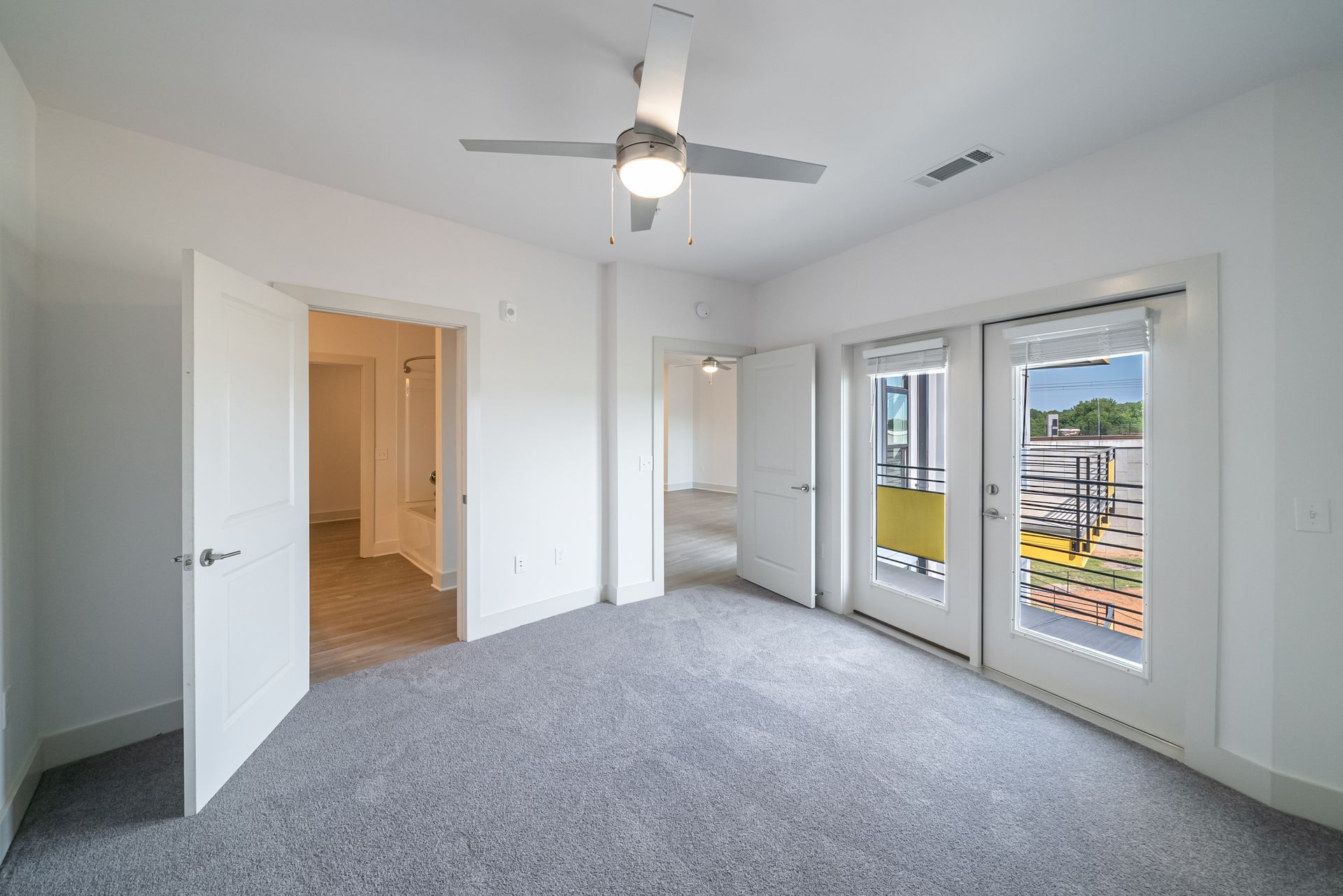 White bedroom with carpet, a ceiling fan, open doors to a hallway and balcony with city view.