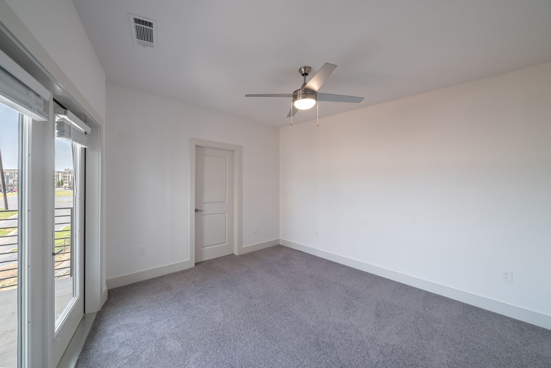 Empty bedroom with gray carpet, white walls, door, ceiling fan, and large windows.