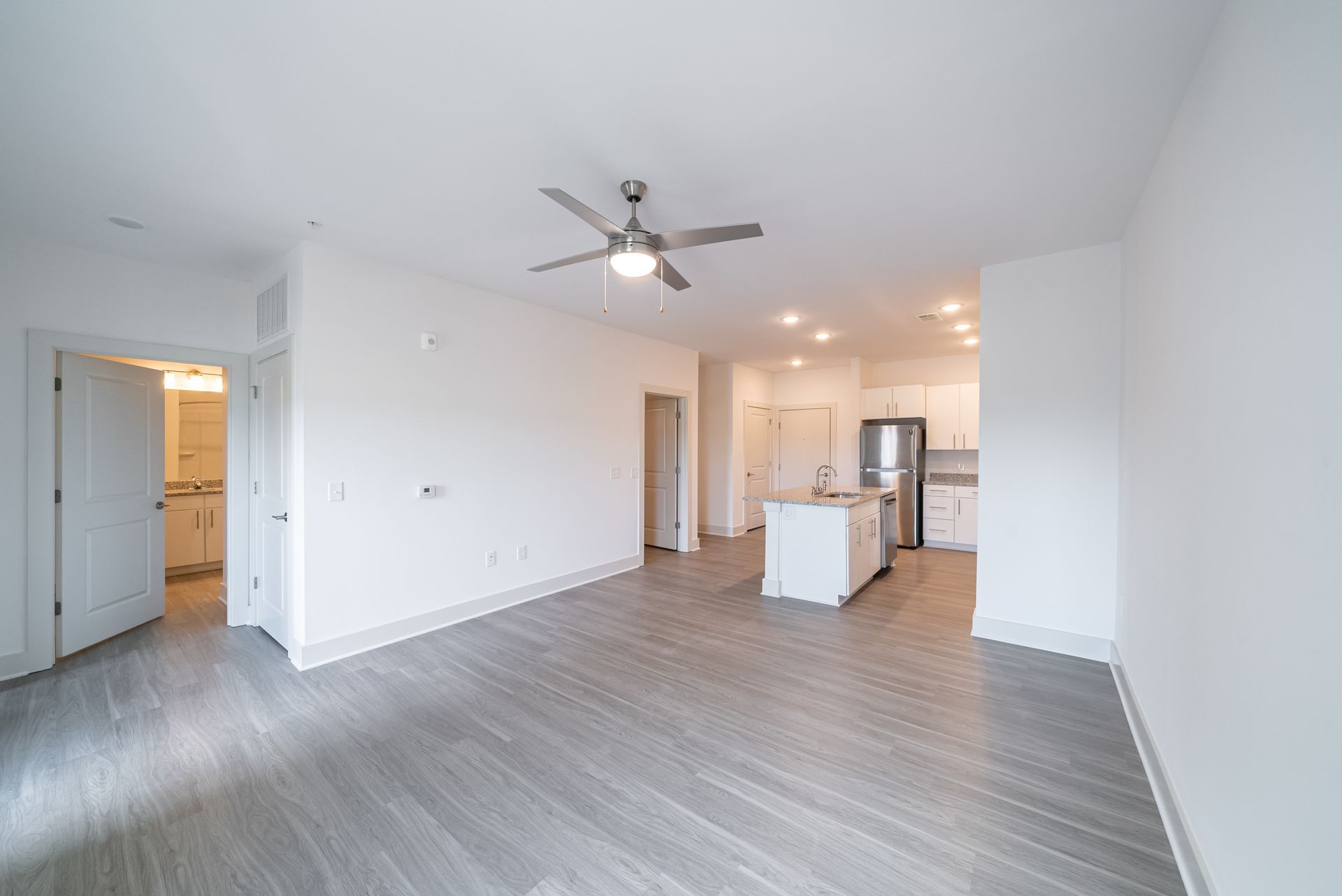 Spacious, empty apartment living area with wood-look flooring, white walls, and a ceiling fan, leading to a kitchen.