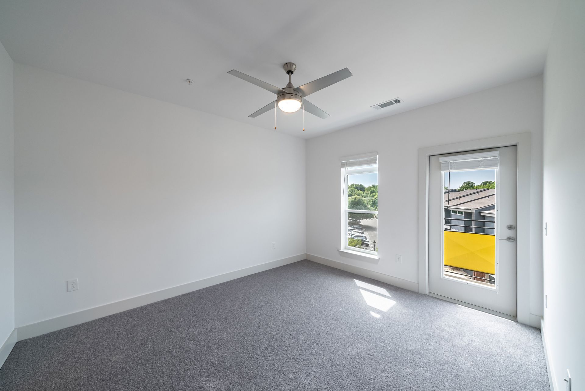 Empty room with gray carpet, white walls, two windows, and a door to a balcony. Ceiling fan is centered.