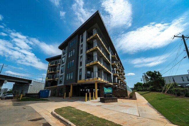 Multi-story apartment building with balconies, grey exterior, near a bridge and green space.