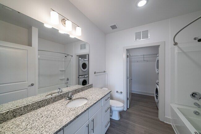 Bathroom with granite countertop, vanity, toilet, shower, and stacked washer/dryer. White and gray color scheme.