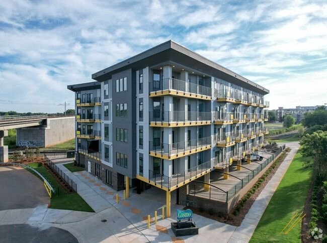 Modern apartment building, gray and yellow exterior, with balconies, against a blue sky, and a nearby bridge.
