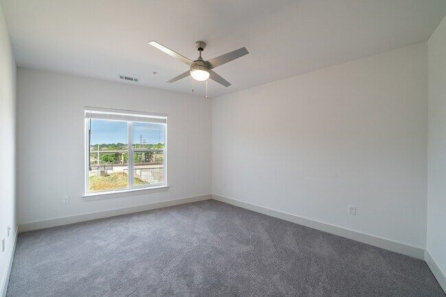 Empty bedroom with gray carpet, white walls, window, and ceiling fan.