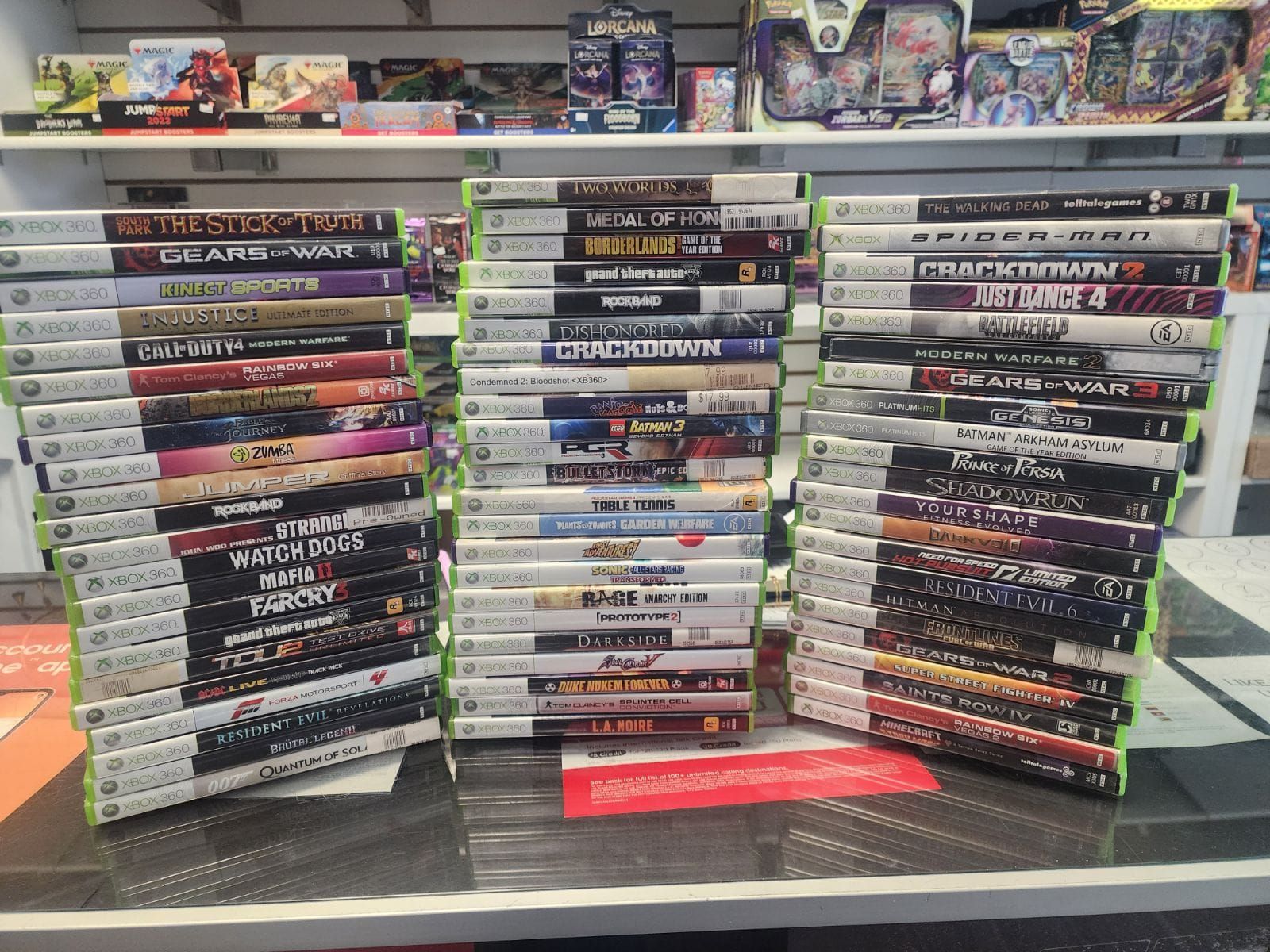 Stacks of Xbox 360 game cases on a counter inside a store, games are displayed by spine. Shelves of other merchandise are in the background.