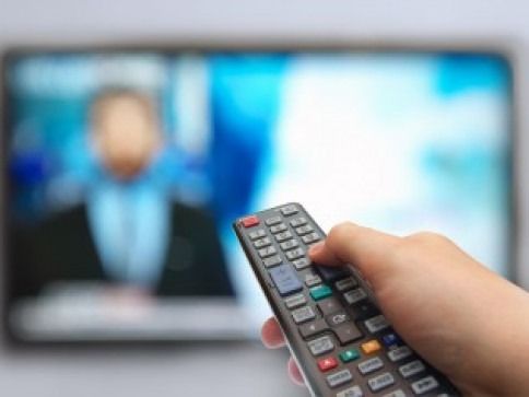 Hand holding a remote control pointing at a blurred television screen.