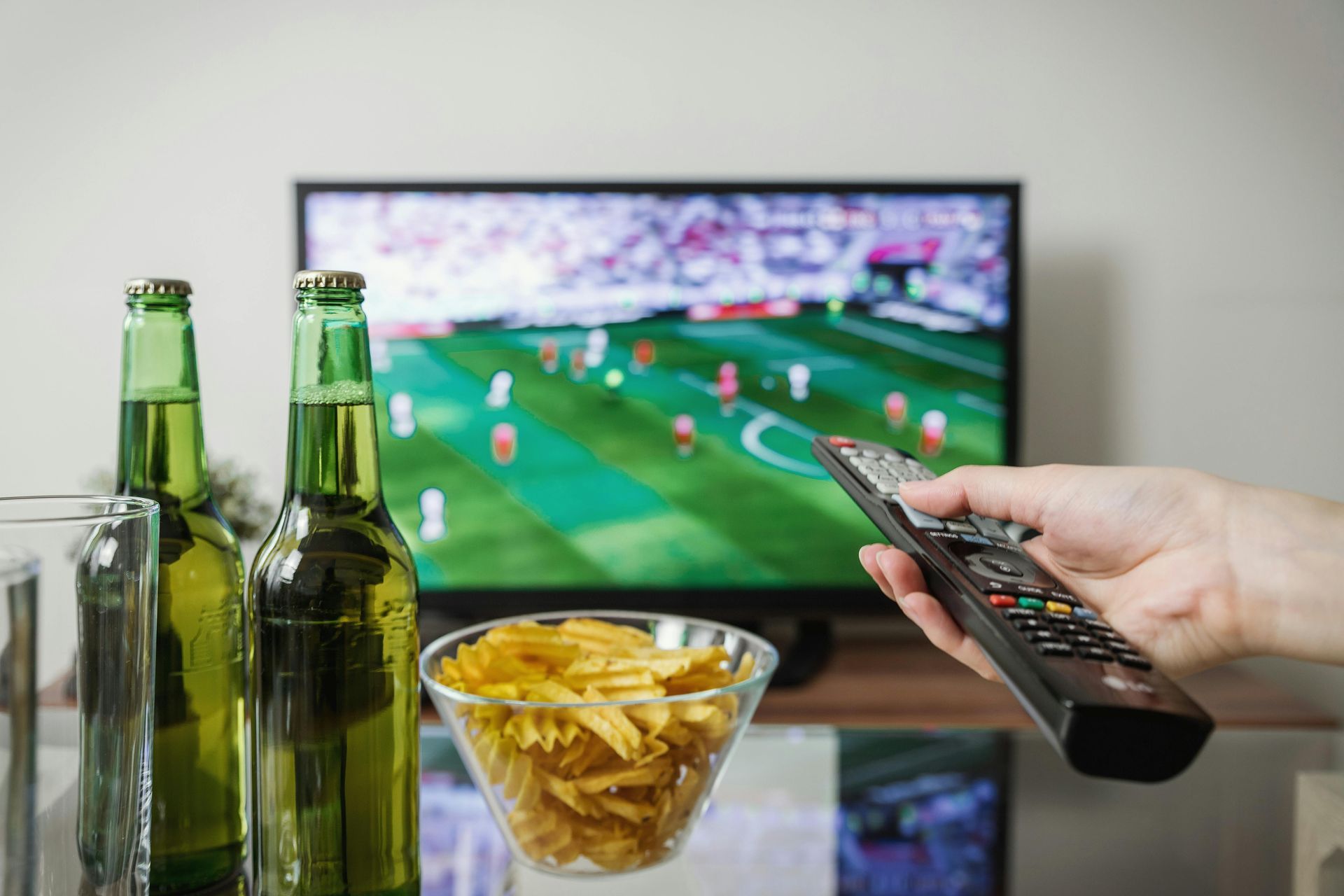 Bottles of beer and bowl of chips sit in front of a TV showing a soccer game; hand holding remote.