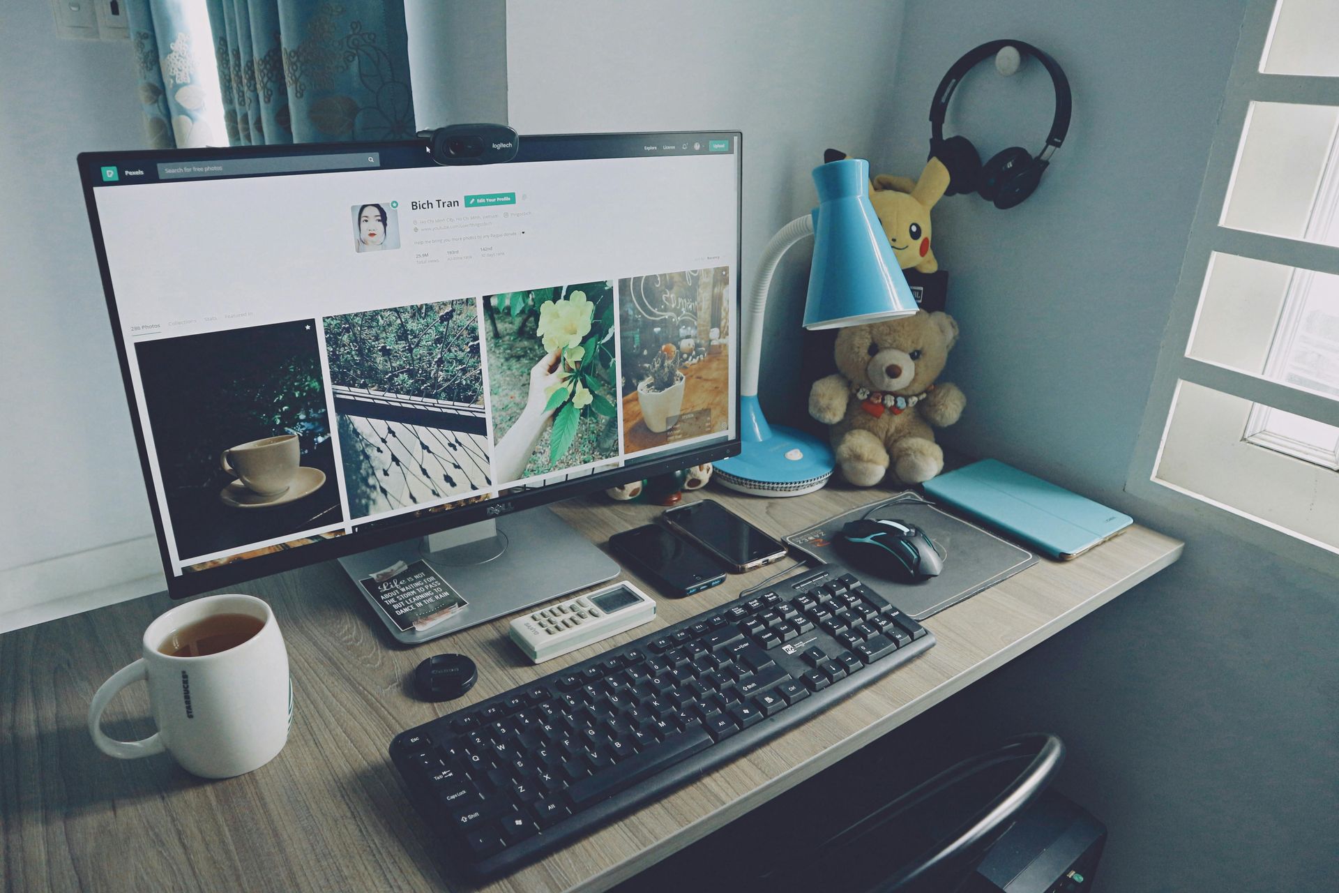 Desk with computer, keyboard, mouse, mug of tea, lamp, teddy bear, and headphones.