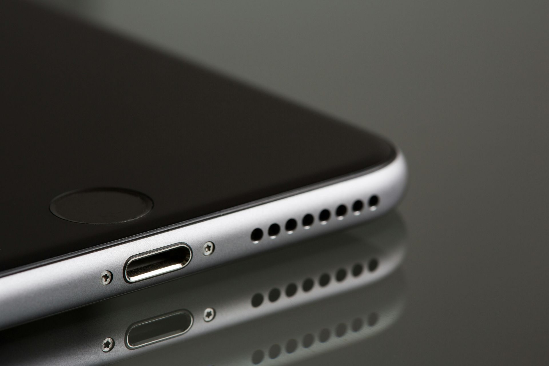 Close-up of a silver smartphone, showing the charging port, speaker holes, and its reflection on a glossy surface.