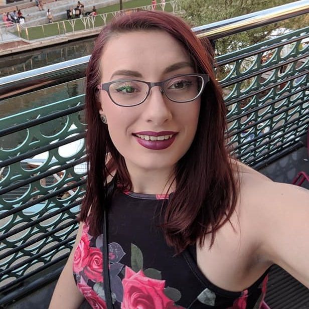 A woman wearing glasses and a floral top is taking a selfie on a balcony.