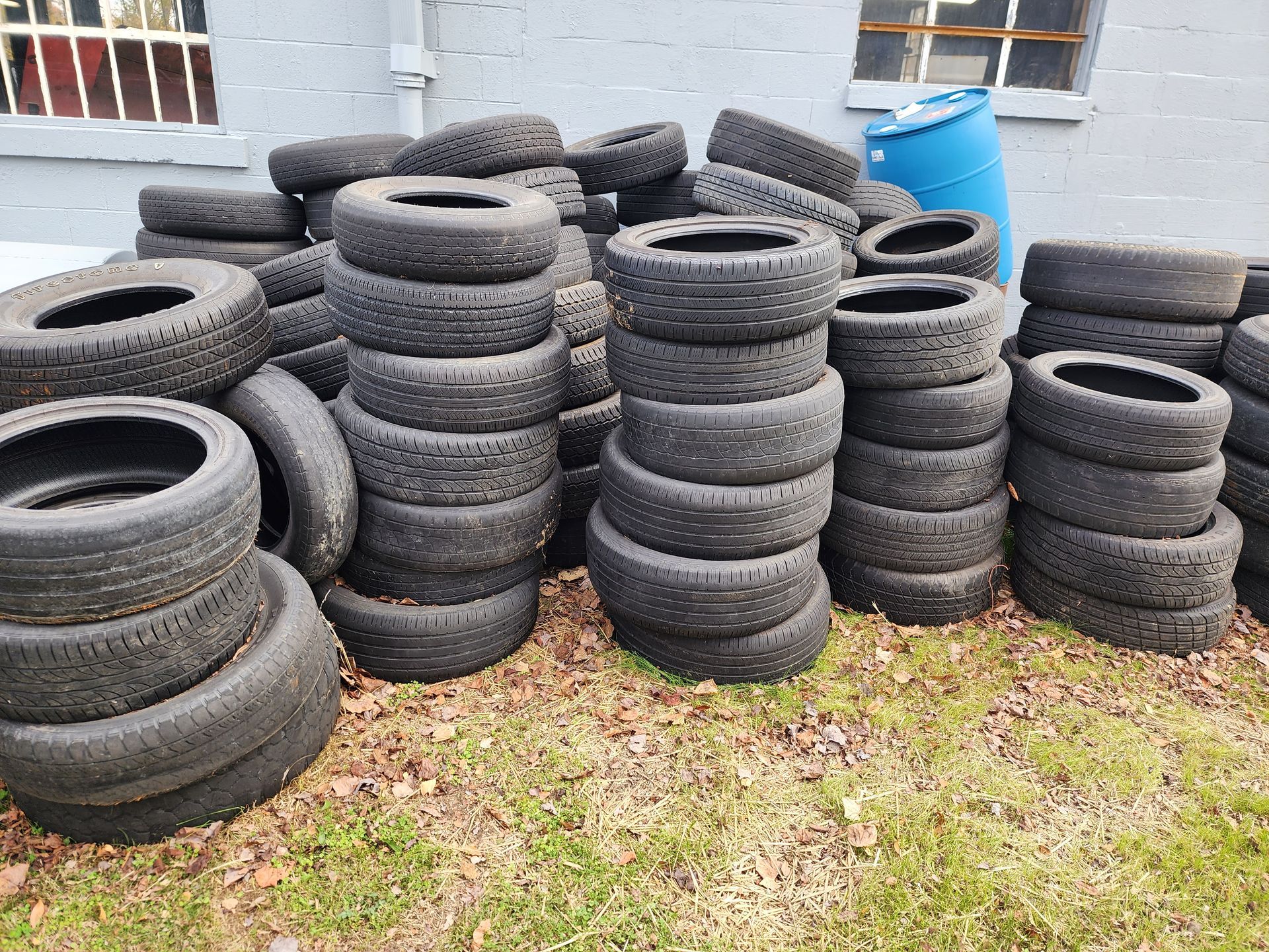Pile of used tires stacked outdoors near a building, with a blue barrel visible.